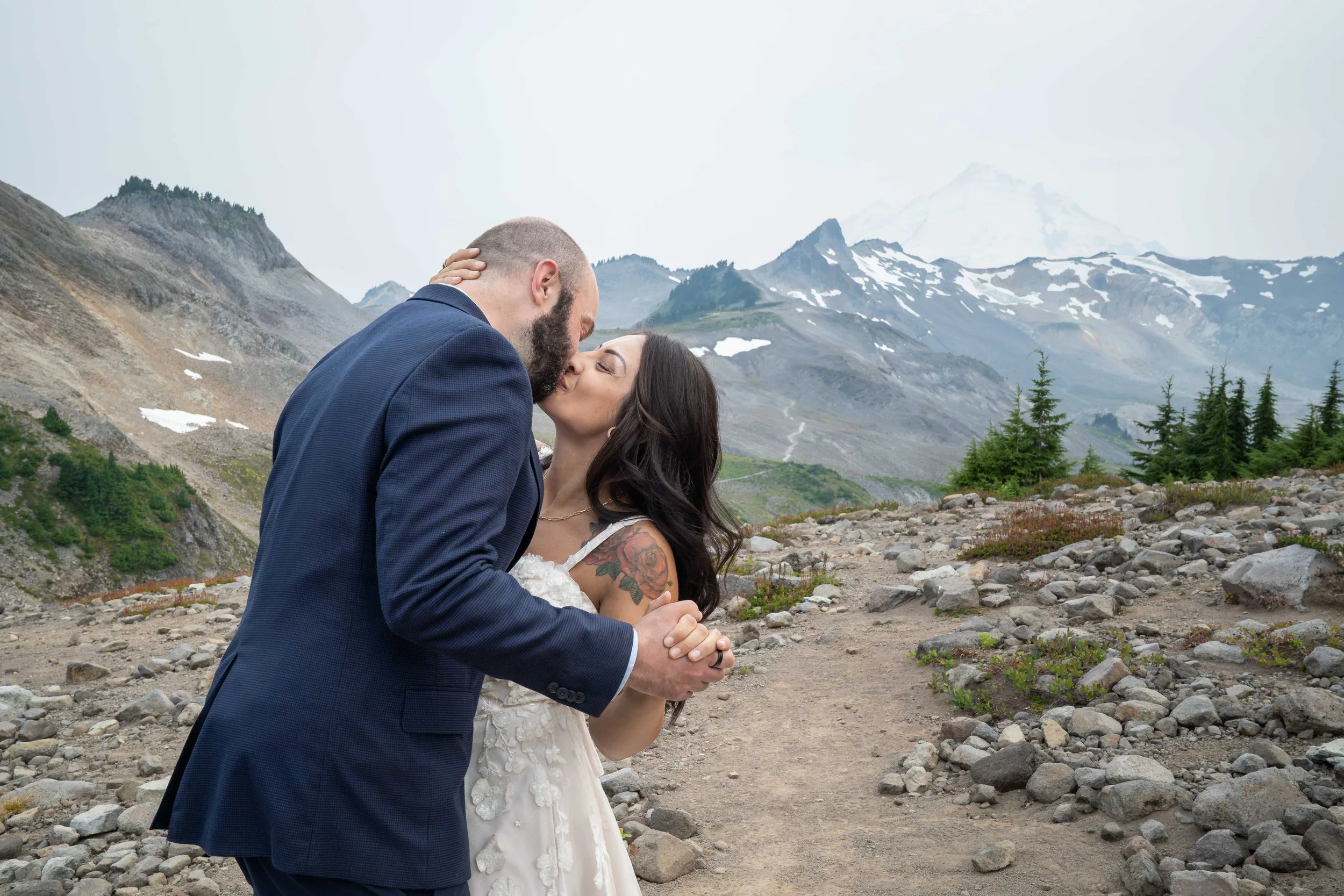 A bride and groom kissing during their first dance on Ptarmigan Ridge as part of their Mt. Baker elopement. The Ptarmigan Ridge trail stretches behind them, with Coleman Pinnacle and the summit of Mt. Baker visible in the background.