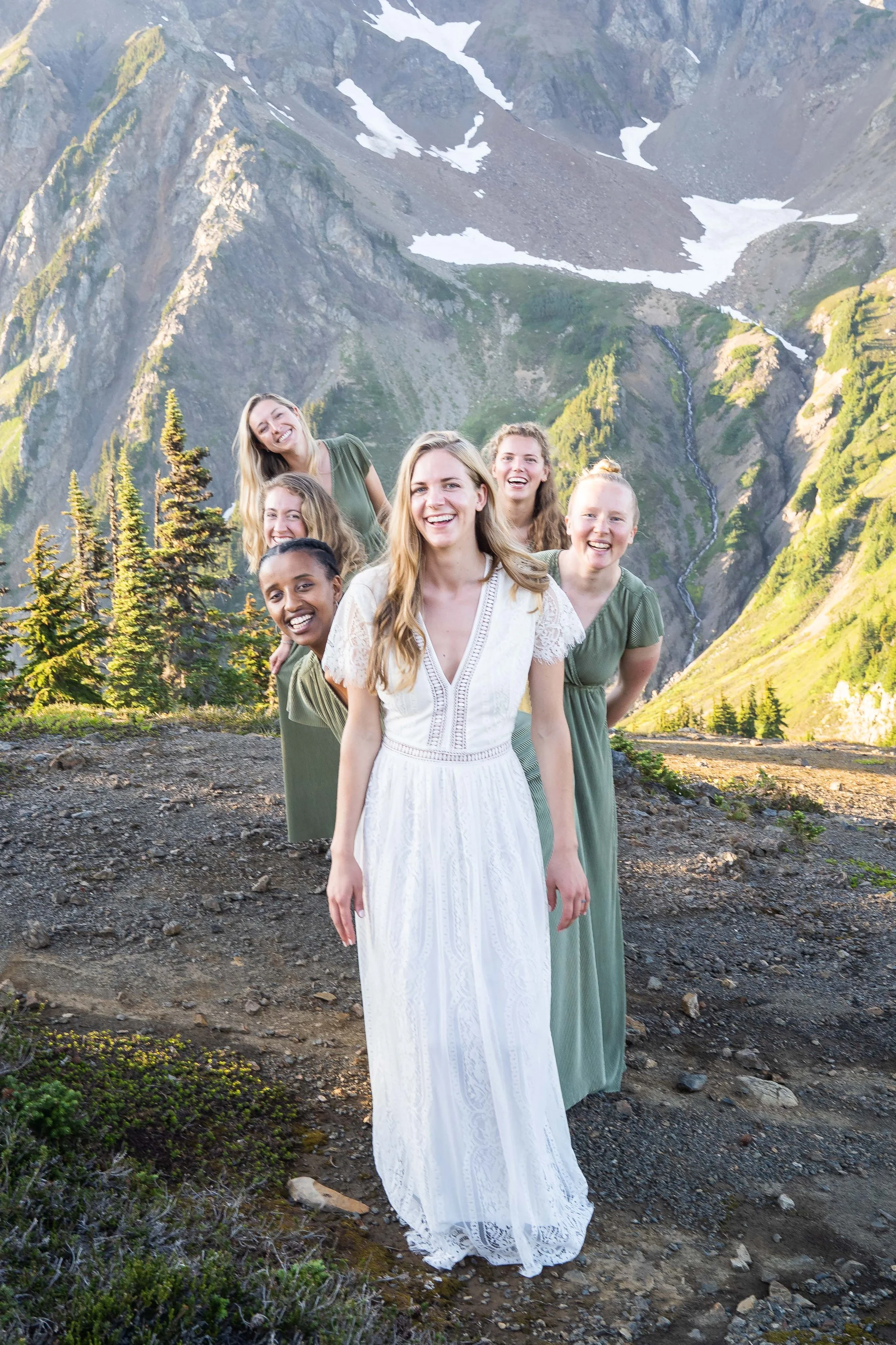 A group photo of a bride in her wedding dress and her bridesmaids in matching green dresses during a vow exchange at High Pass in the North Cascades, Washington. The bridesmaids are arranged so that their heads are sticking out behind the bride.