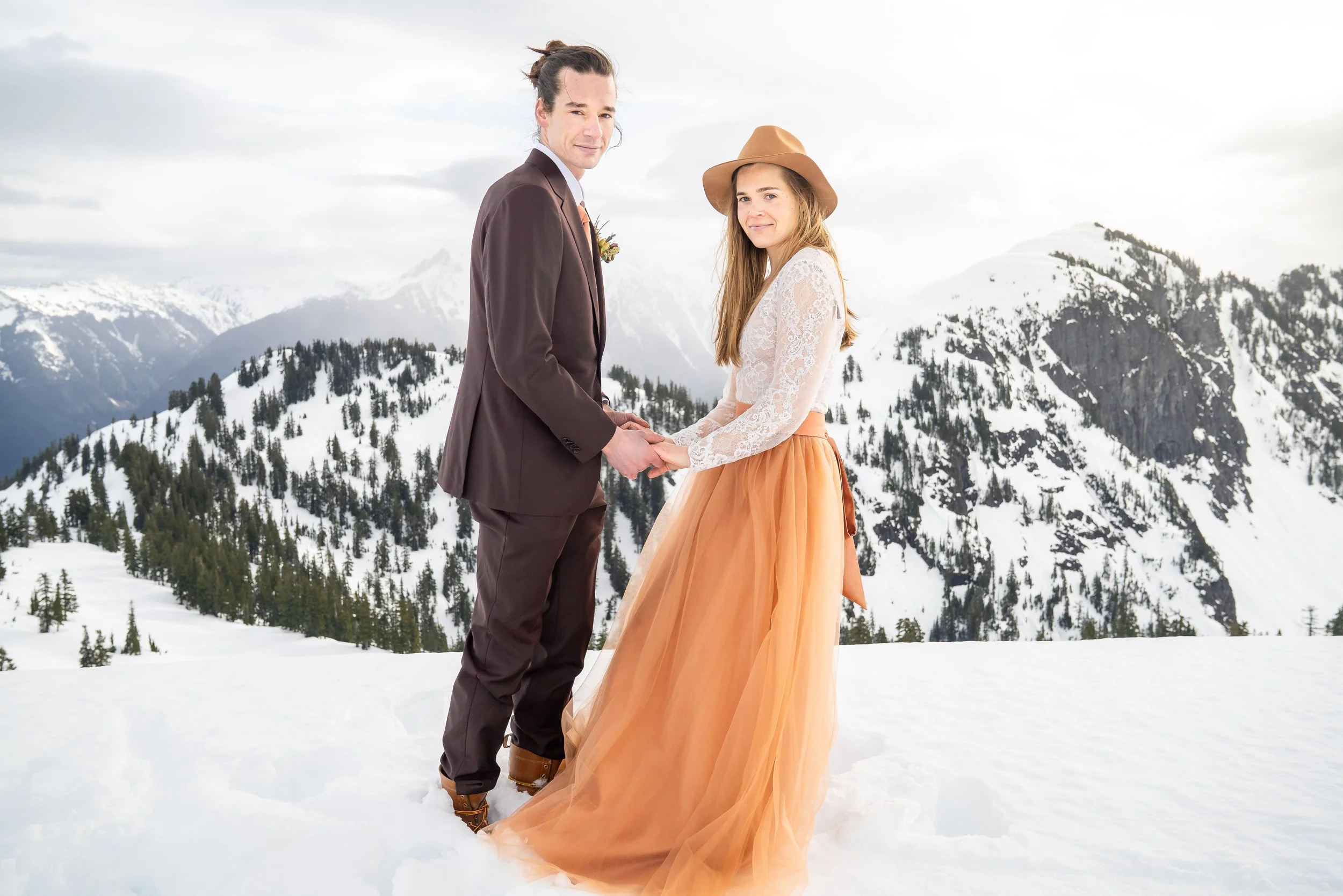 A photo of a bride and groom in wedding clothes standing together and holding hands. The groom is in a brown suit and the bride is wearing an orange and white wedding dress. They're standing in the snow at Artist Point with mountains behind them.