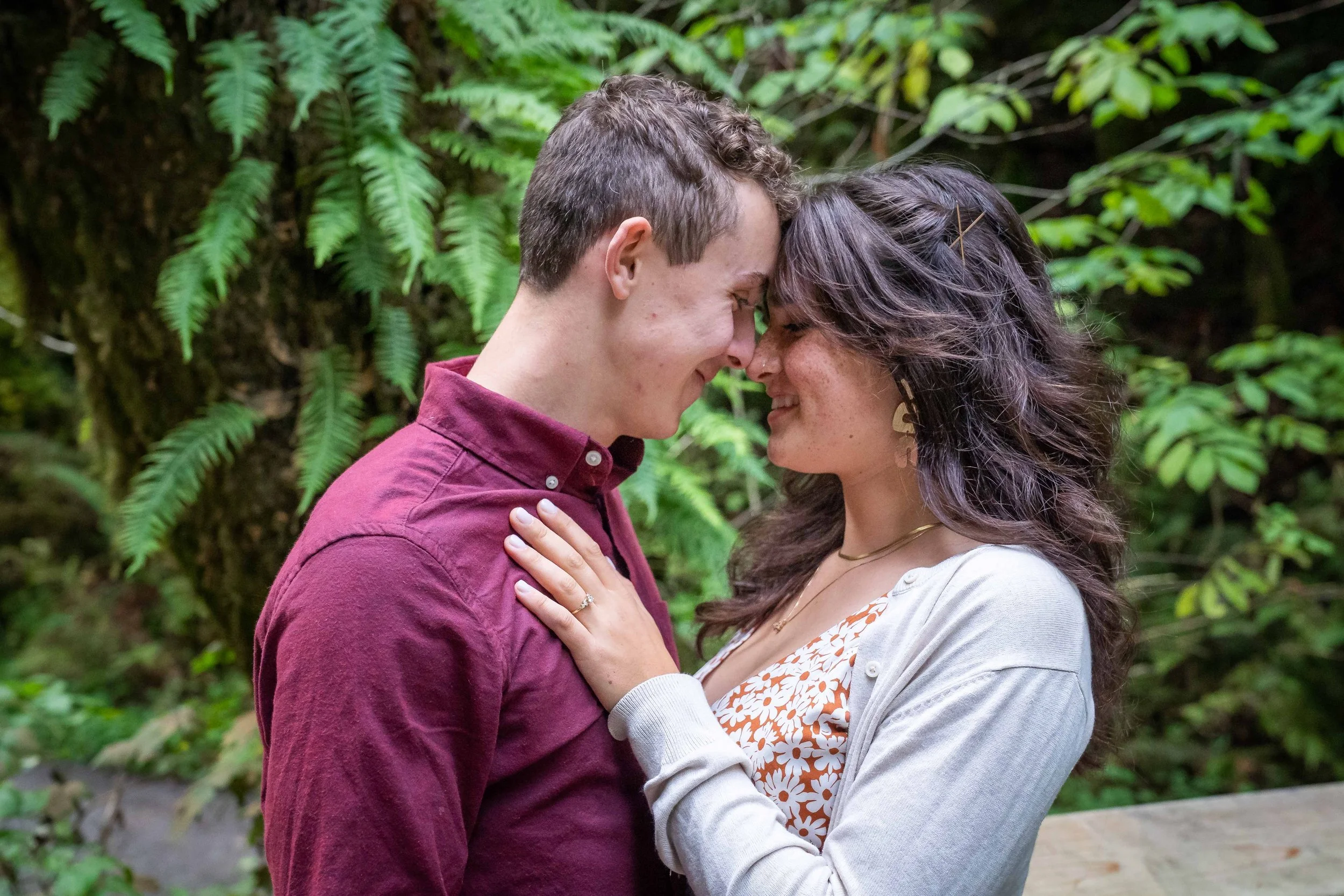 A photo of a couple in a button-down shirt and dress standing together and touching foreheads, with the woman's hand on the man's chest. The woman's engagement ring is clearly visible and there's a lush green forest behind them.