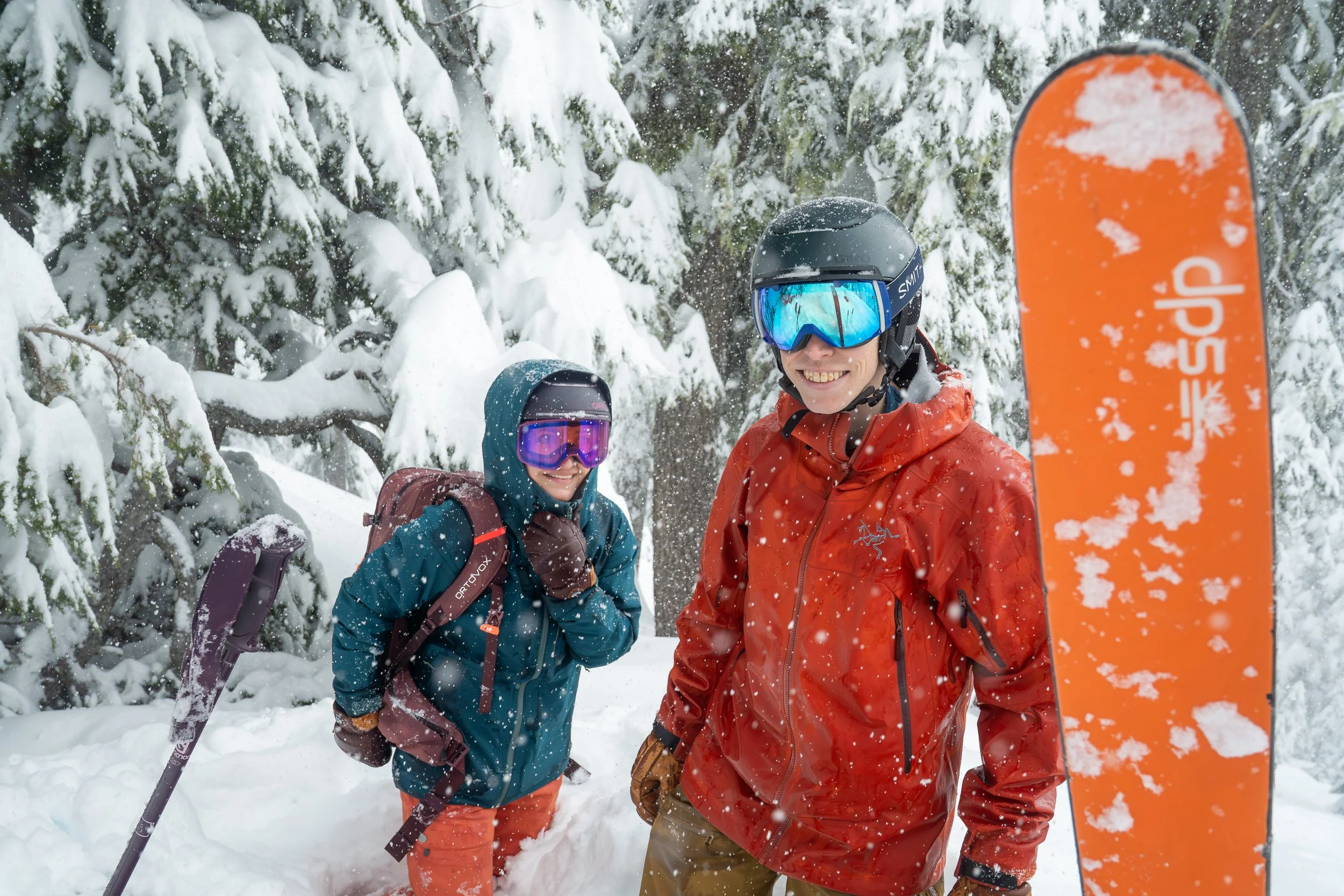 Photo of a couple standing in the snow in a snowy forest as they transition from uphill skinning to downhill skiing. A ski is upright in the foreground and the man and woman are standing together smiling at the camera.