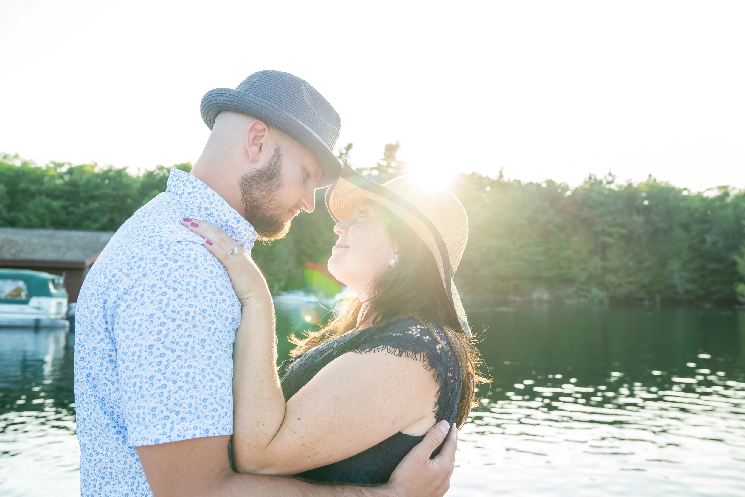A photo of a couple holding each other and looking into each other's eyes. There's a lake behind them and a glowing sunset.