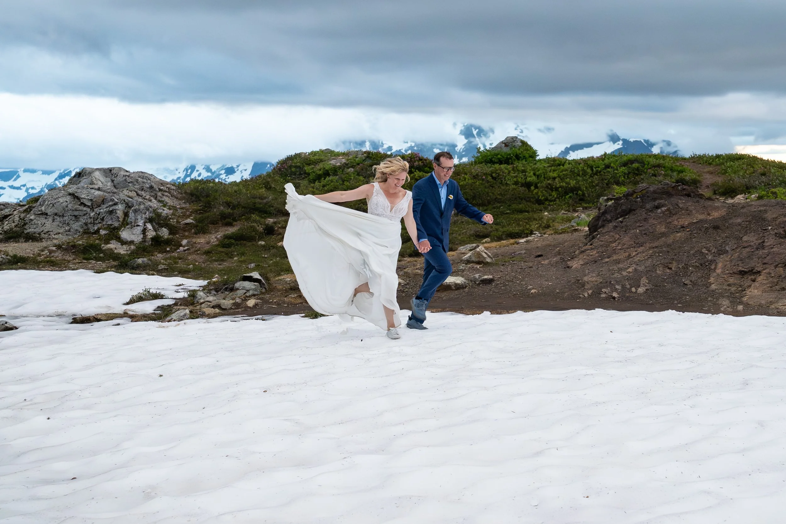 A bride and groom in wedding clothes running through snow near Yellow Aster Butte in the North Cascades after their elopement. Mt. Shuksan is partially visible in a cloud behind them.