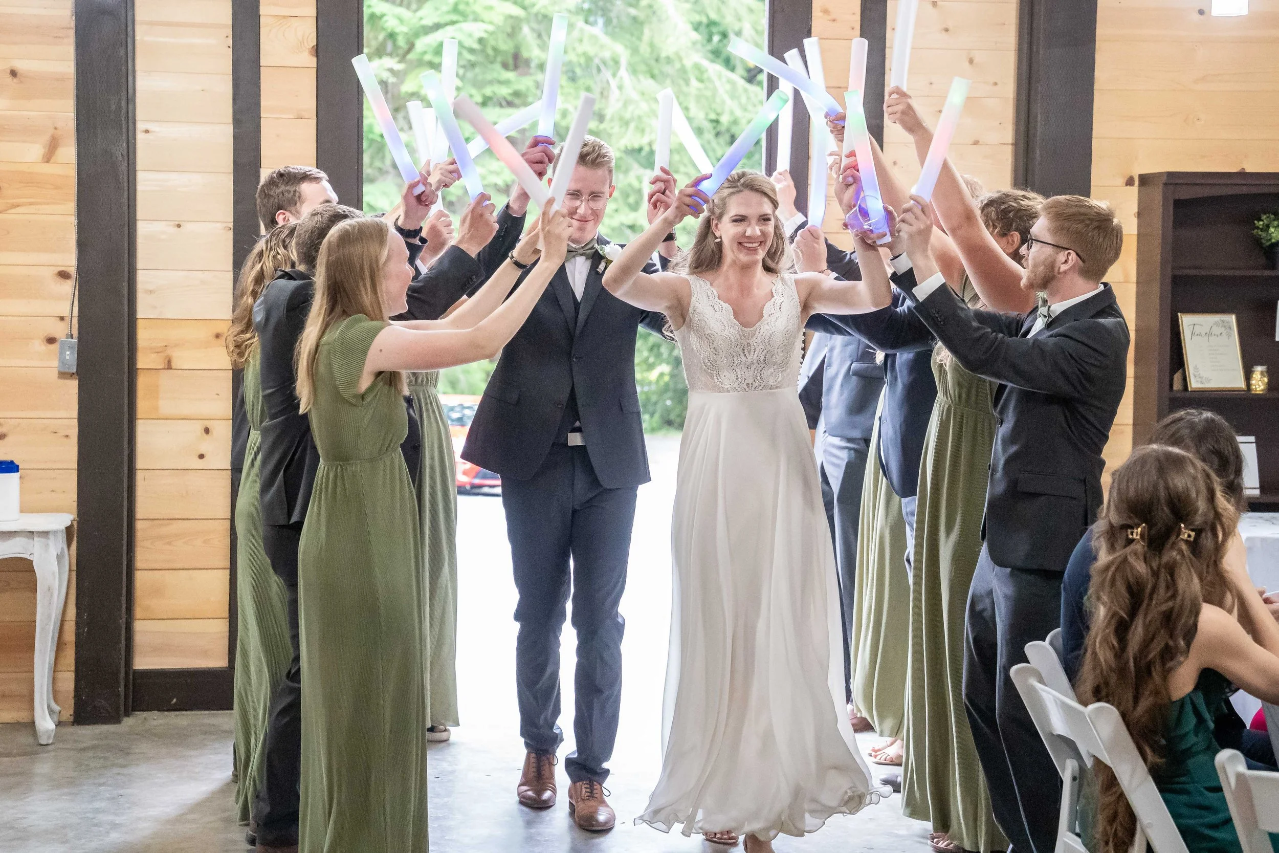 A bride and groom's grand entrance to their wedding reception in a bright, wooden venue. The bride and groom are holding glowsticks and are walking through a tunnel of glowsticks held by the bridesmaids and groomsmen.