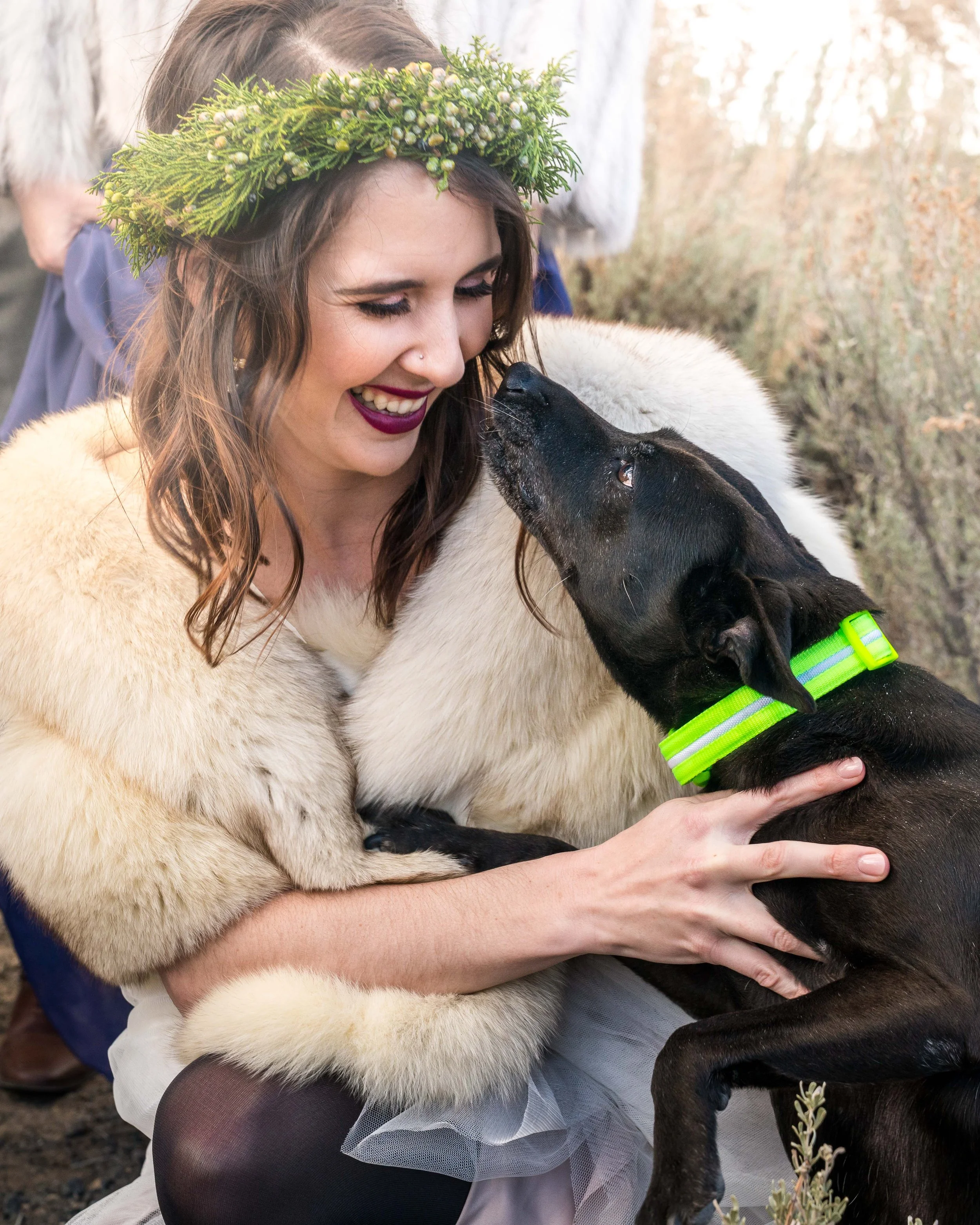 A photo of a bride in a wedding dress and white fur coat smiling and holding a black dog.