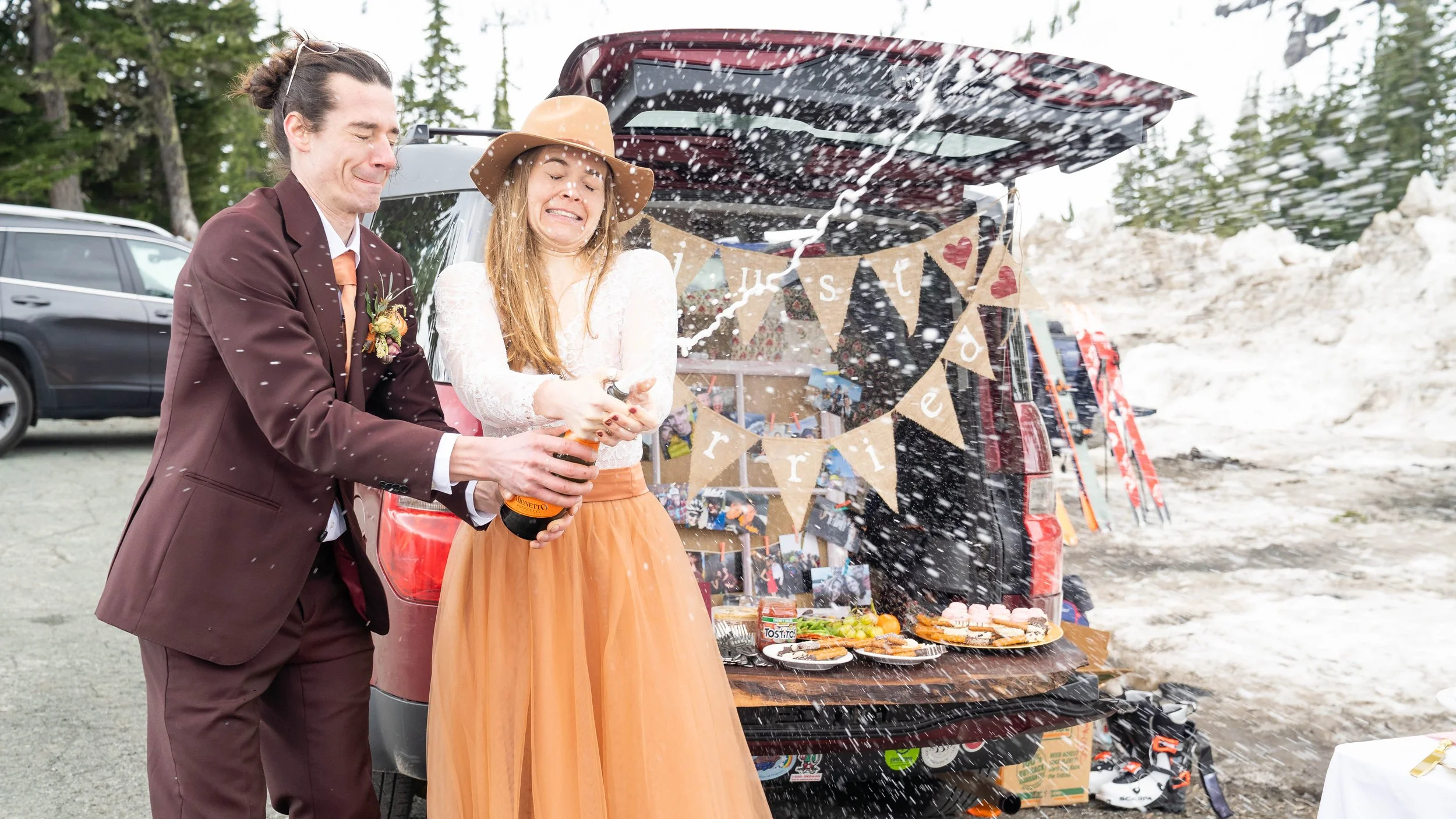 A bride and groom popping champagne during a tailgate reception party at the Heather Meadows parking lot after a backcountry ski elopement at Artist Point. They are standing in front of a car's open trunk that has snacks and a "Just Married" sign.