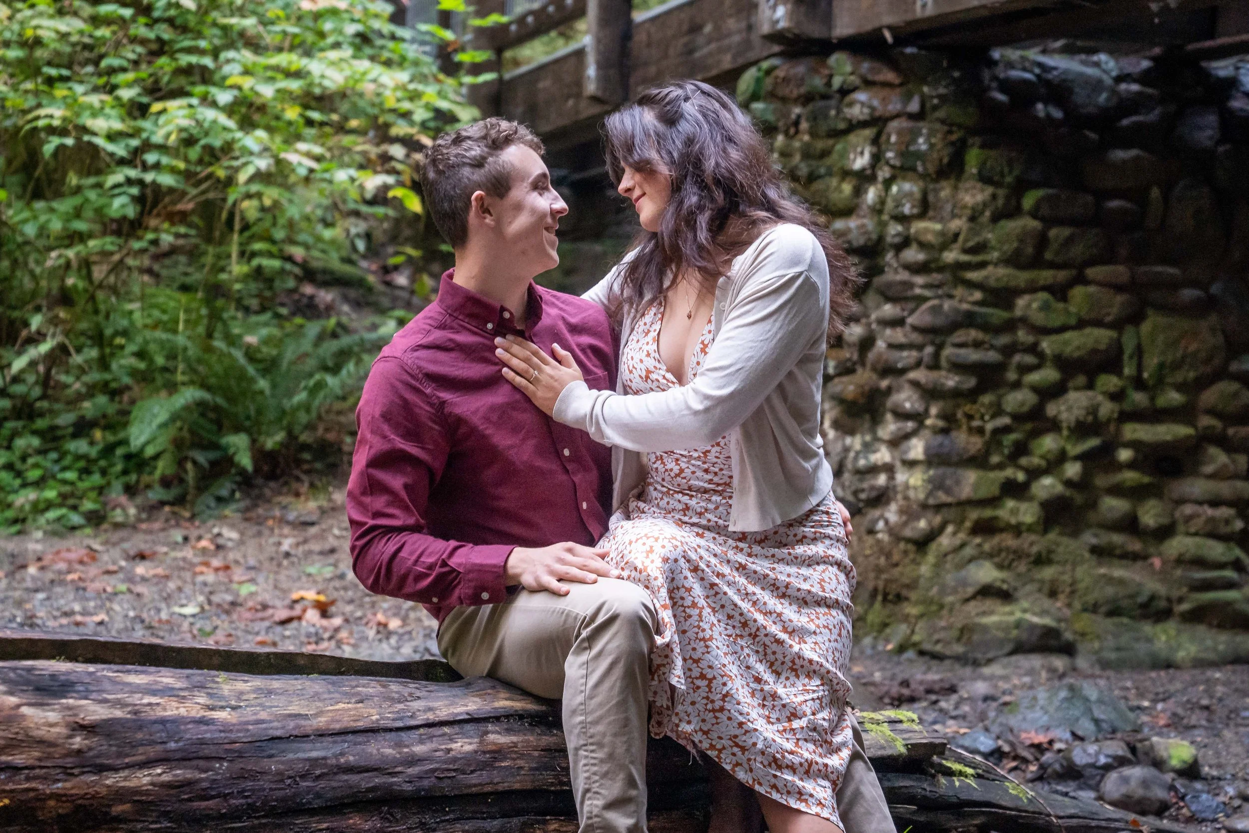 A photo of a young couple sitting together on a log in Arroyo Park. There's a stone bridge behind them and the woman is sitting on the man's leg with a hand on his chest.