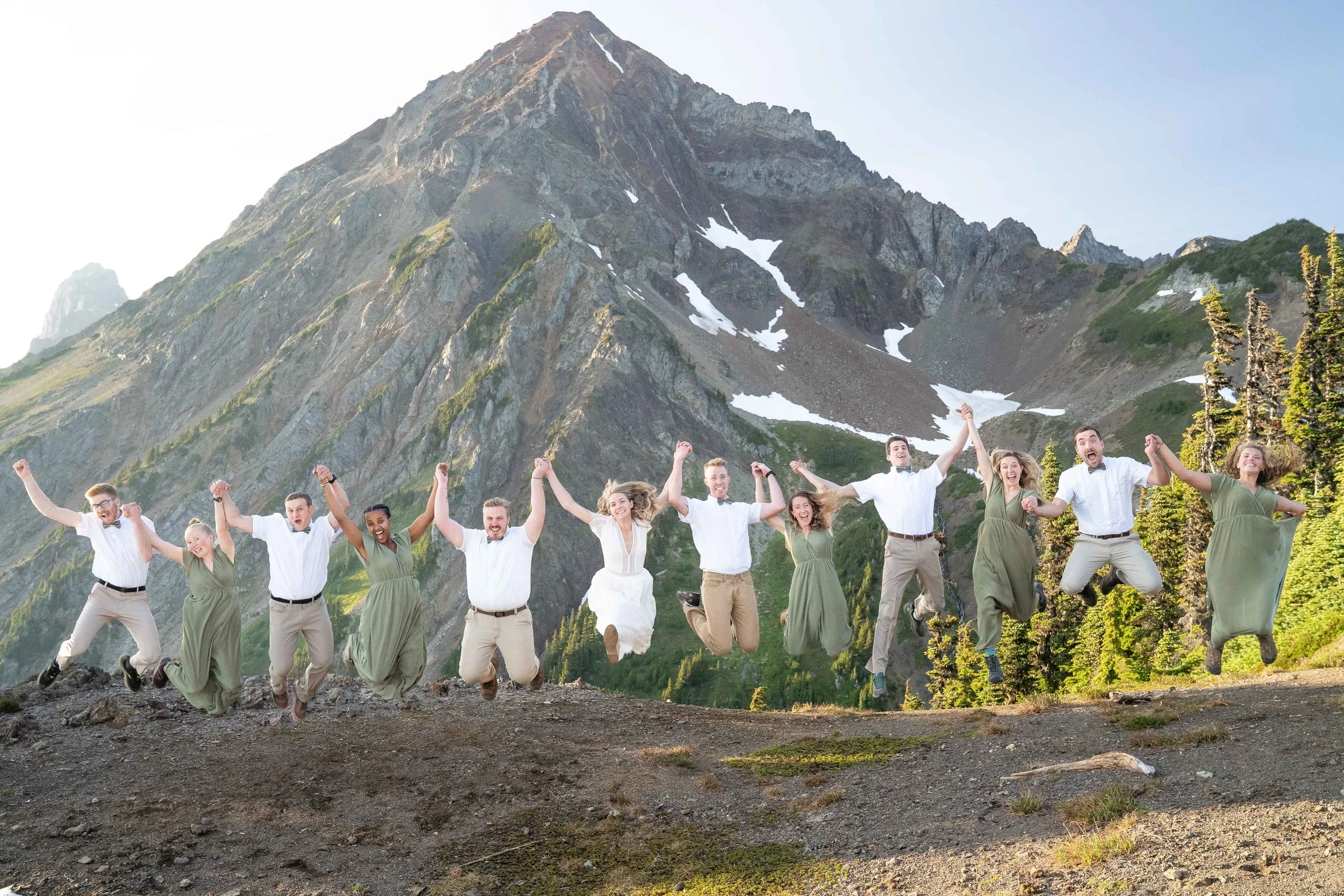 A full bridal party including bridesmaids and groomsmen holding hands and jumping in the air in front of Mt. Larrabee, near High Pass in the North Cascades, at sunset. Each person in the group is wearing wedding clothes and hiking boots.
