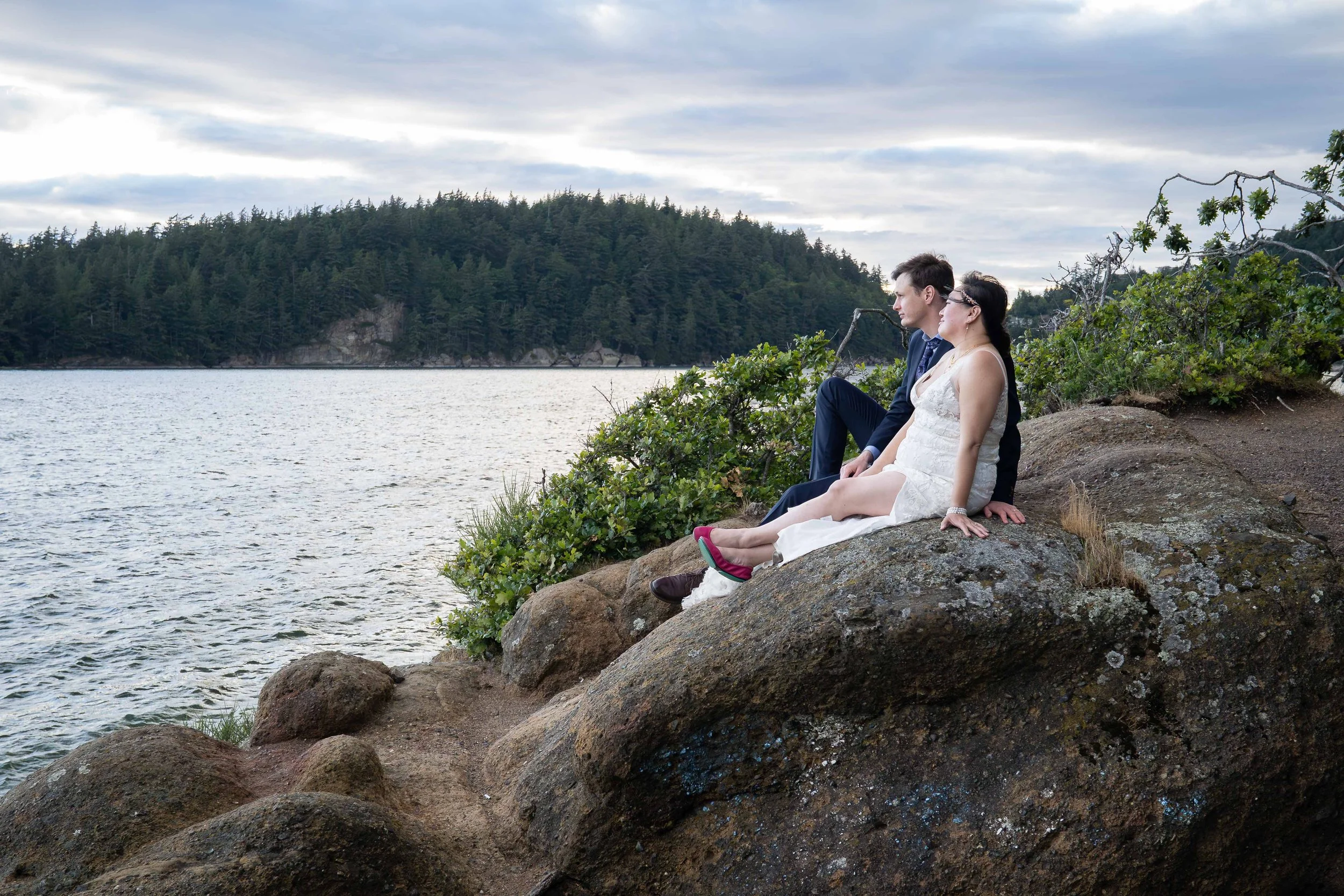 A bride and groom sit together on a rocky bluff at Teddy Bear Cove, in Bellingham, Washington, at sunset. They are relaxing and looking out over the water after eloping in Larrabee State Park earlier in the day.