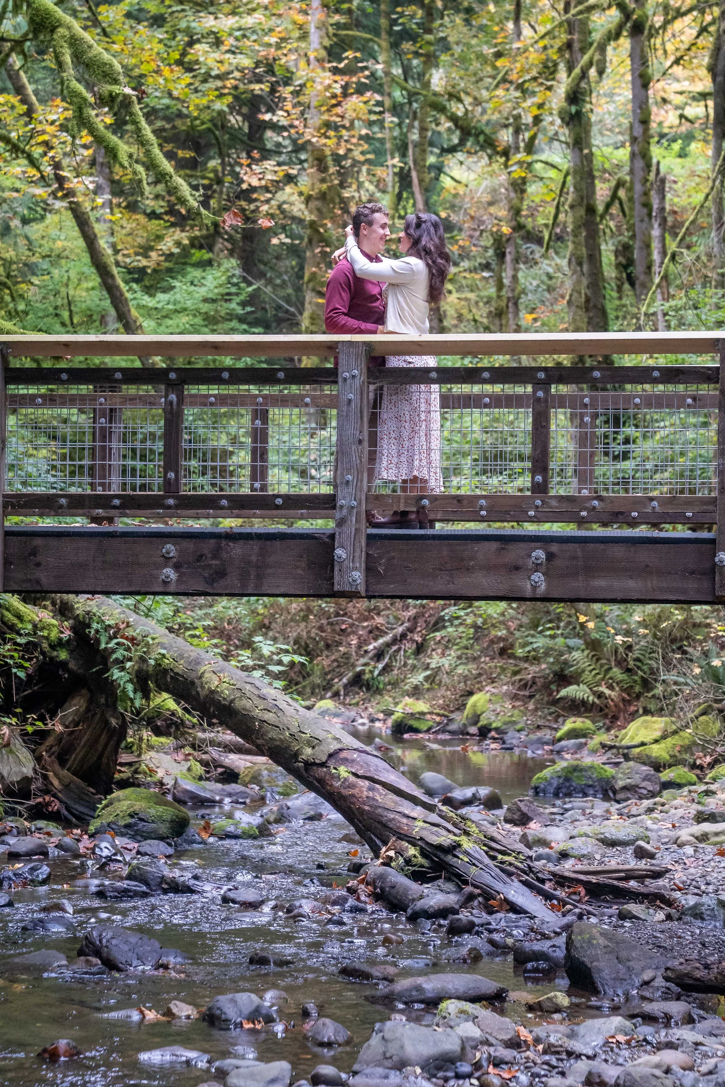 A photo of a couple standing on a wooden bridge in Arroyo Park in Bellingham. They're standing together with the woman's arms around the man's shoulders. There's a creek in the foreground and a mossy forest in the background.