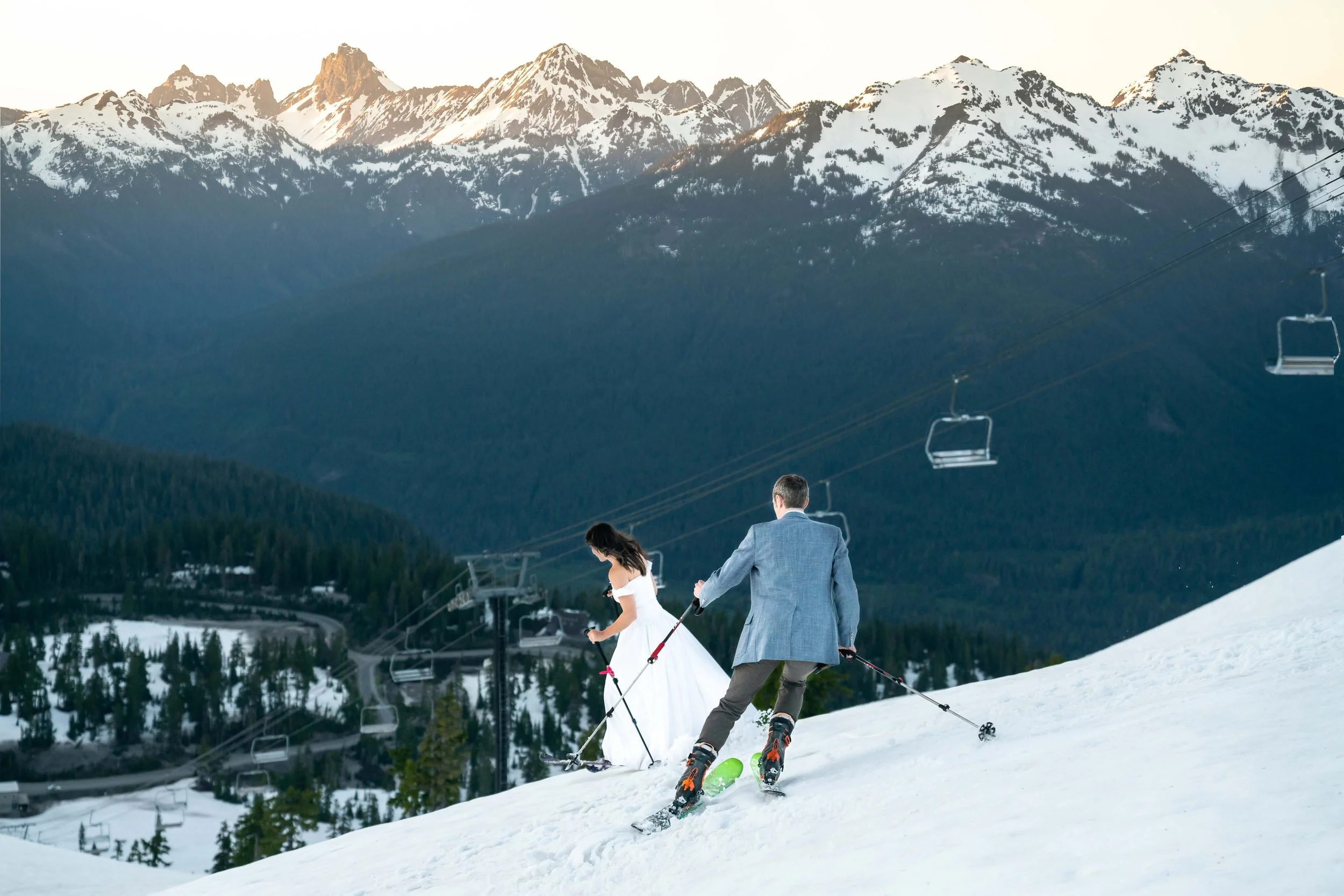 A photo of a bride in a wedding dress and a groom in a suit skiing downhill at Mt. Baker Ski Area, with large snowy mountains in the background.