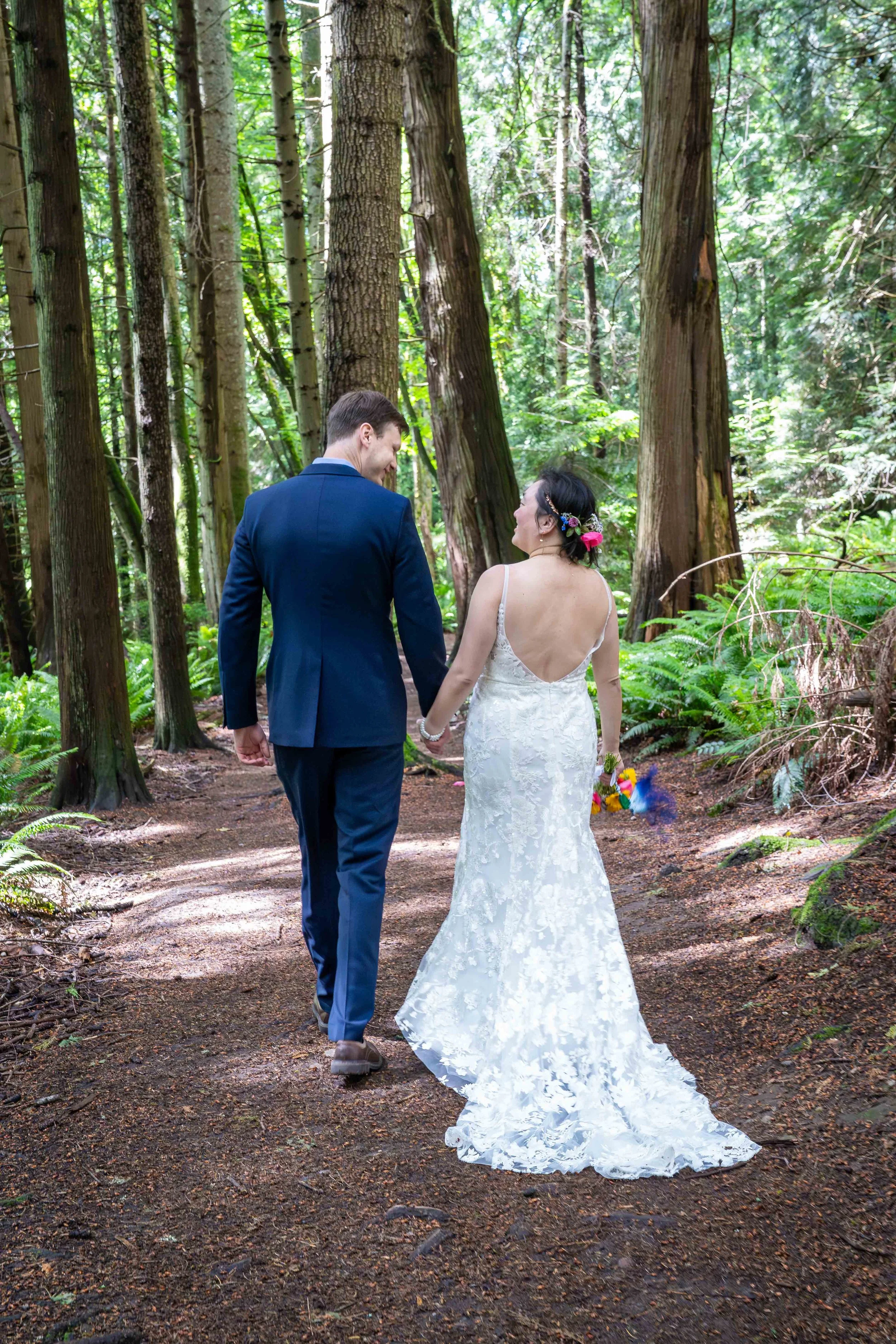 A bride and groom hold hands and walk down the Fragrance Lake Trail after their Larrabee State Park elopement. They are smiling at one another and walking towards a grove of old growth trees and ferns.