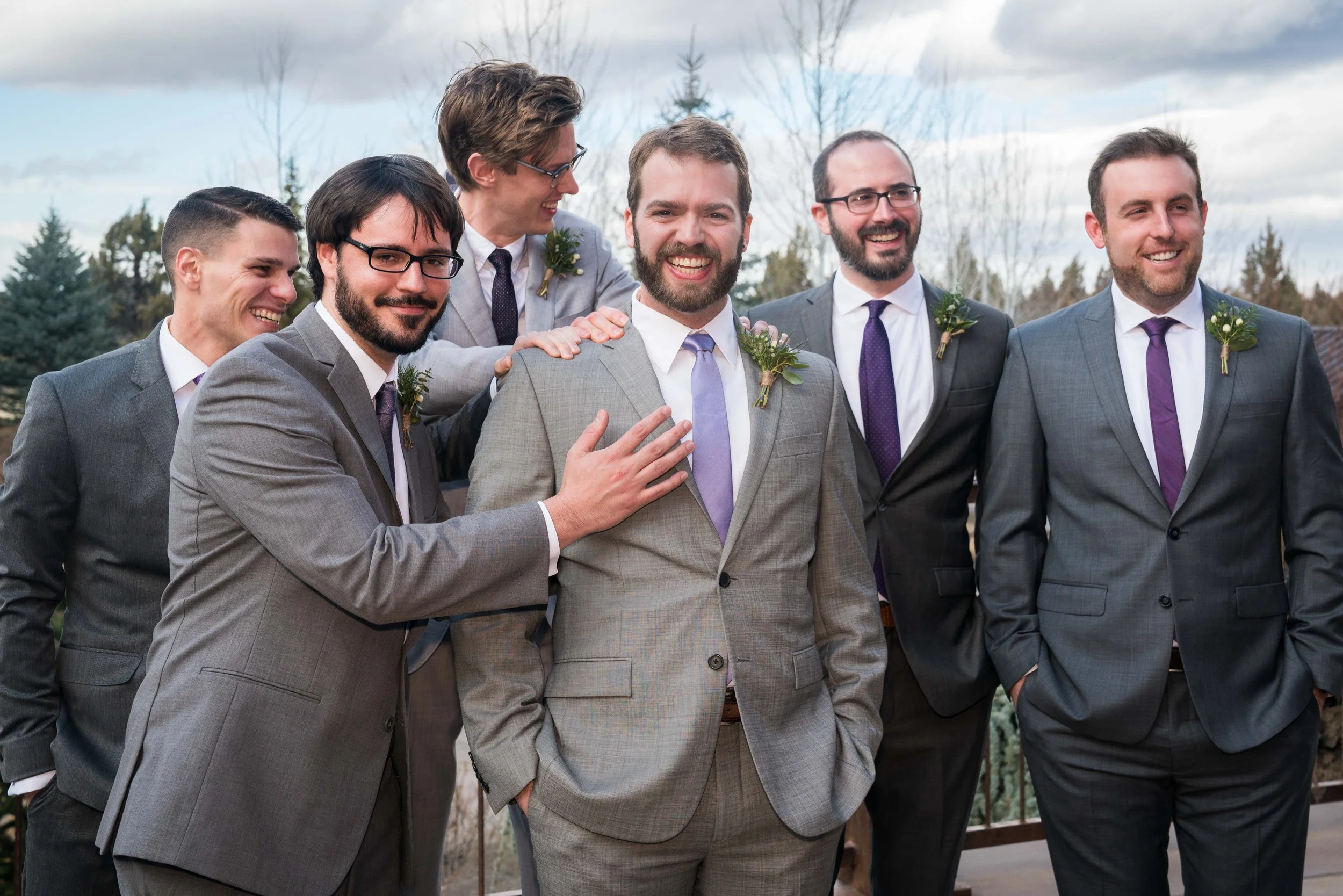 A photo of a groom and groomsmen in suits. The groomsmen are standing arranged around the groom and his best man is patting him on the shoulder.