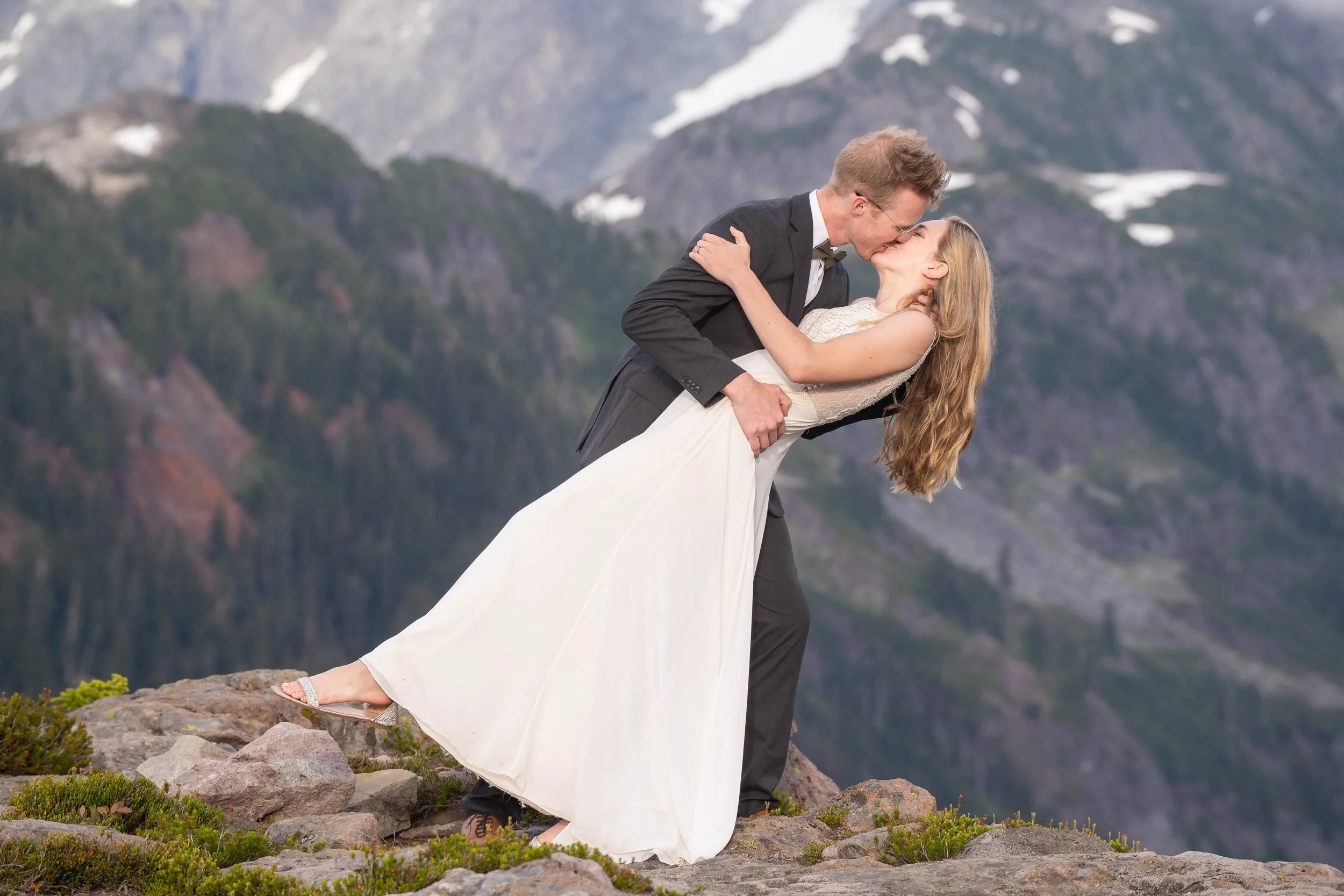 Photo of a bride in a wedding dress and a groom in a suit kissing during an Artist Point elopement. They're standing in a rocky alpine meadow with snowy mountains behind them and the bride is leaned backwards with her dress and sandal in the air.