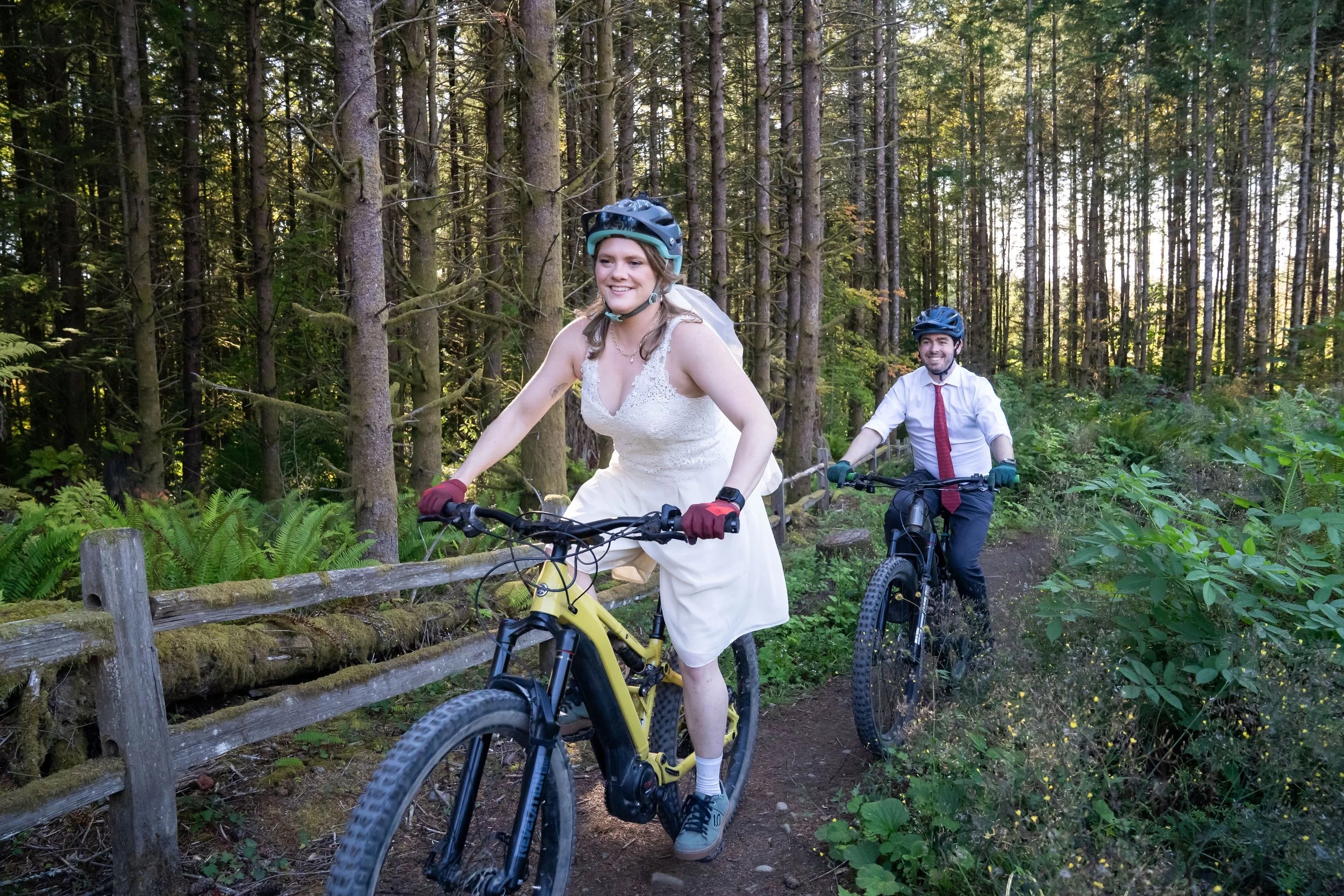 A bride and groom in wedding clothes riding along a fence on the trail New Issues on Galbraith Mountain during their mountain biking elopement.
