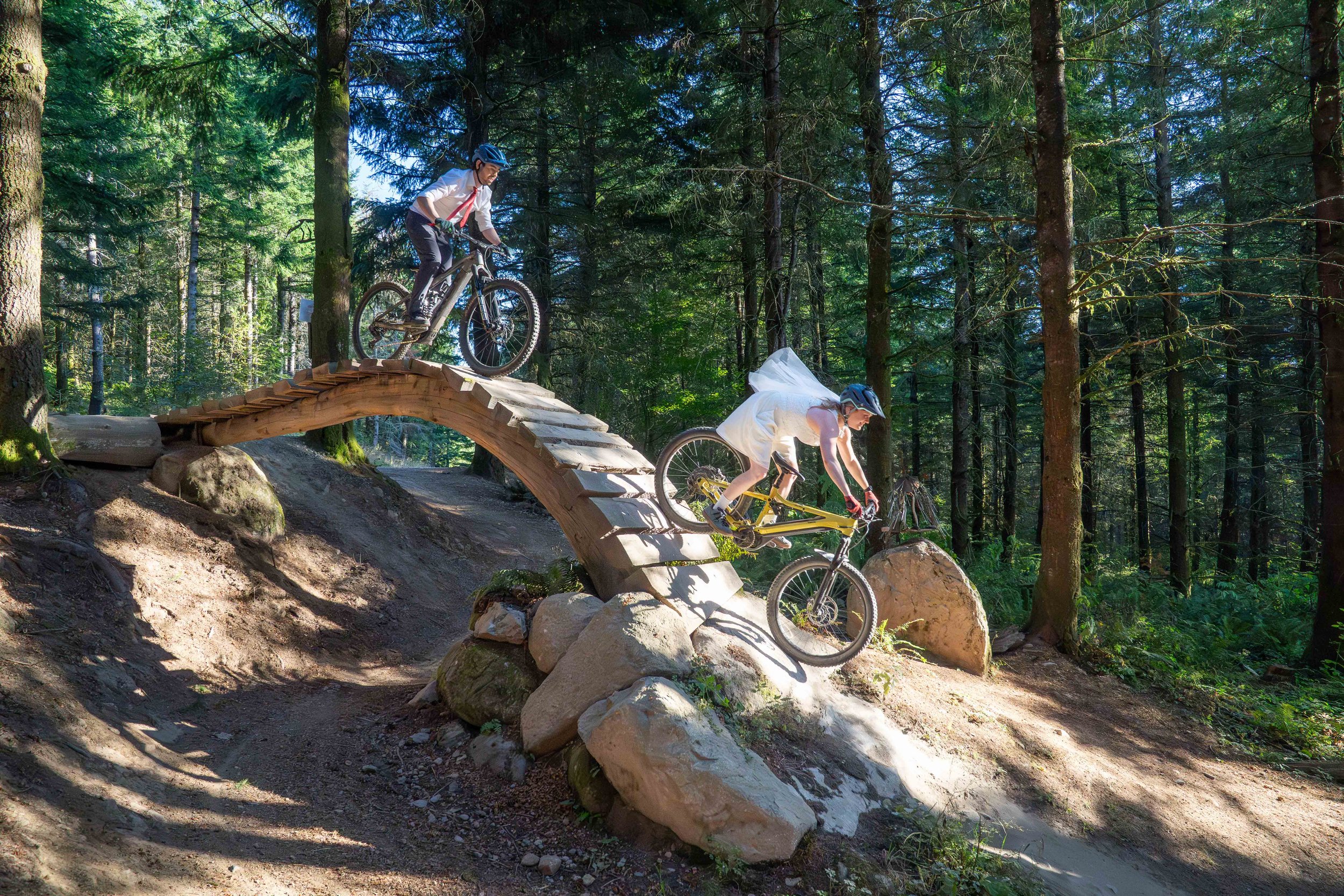 A bride and groom in full wedding clothes riding the Unemployment Line wooden roll simultaneously during their mountain biking elopement photoshoot on Galbraith Mountain, in Bellingham, Washington. The bride is near the bottom of the roll.