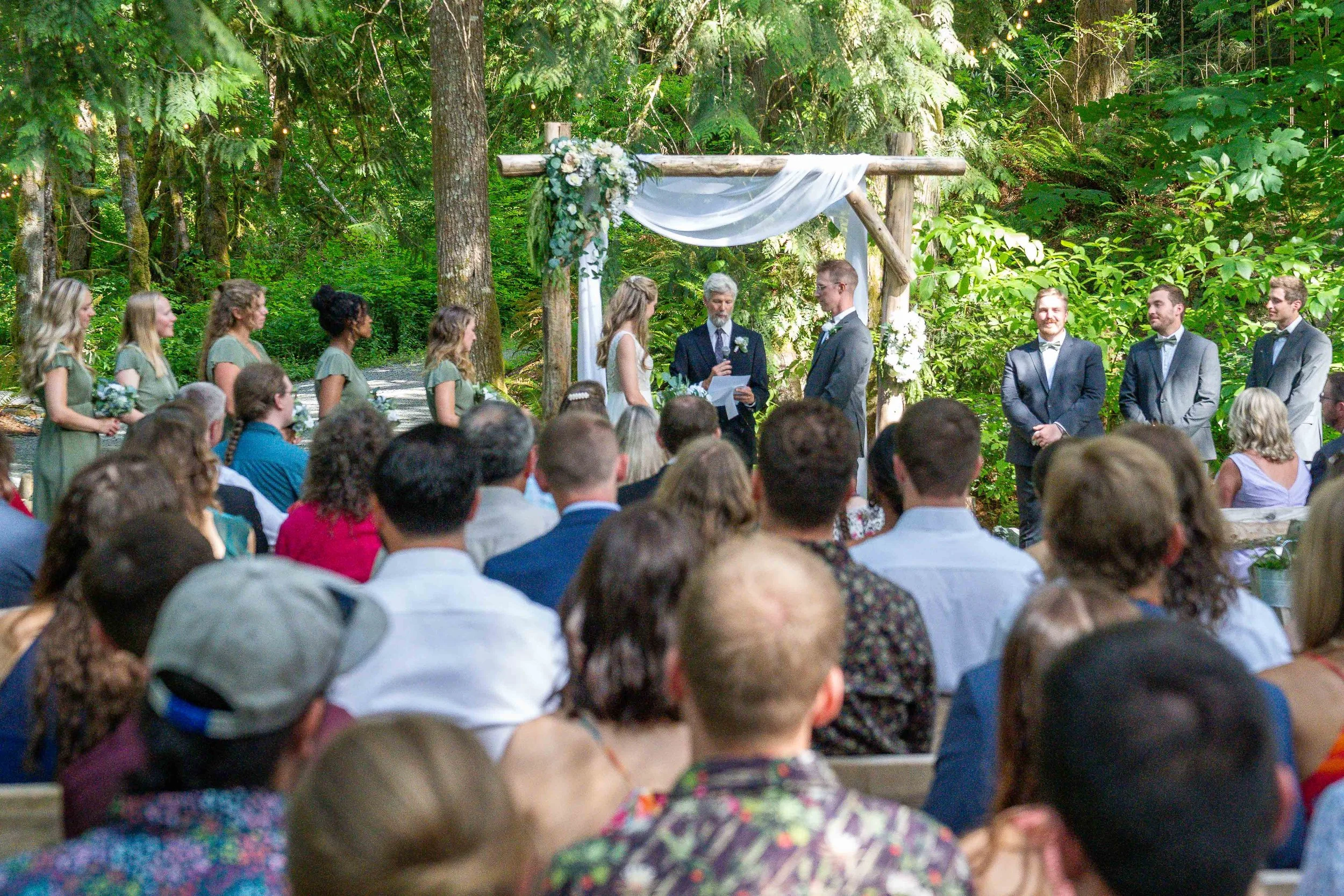 Photo of a forest wedding ceremony at Creekside Celebrations in Skagit County, Washington. The photo shows the bride and groom at the altar with a crowd in front of them and their bridesmaids and groomsmen to their sides.