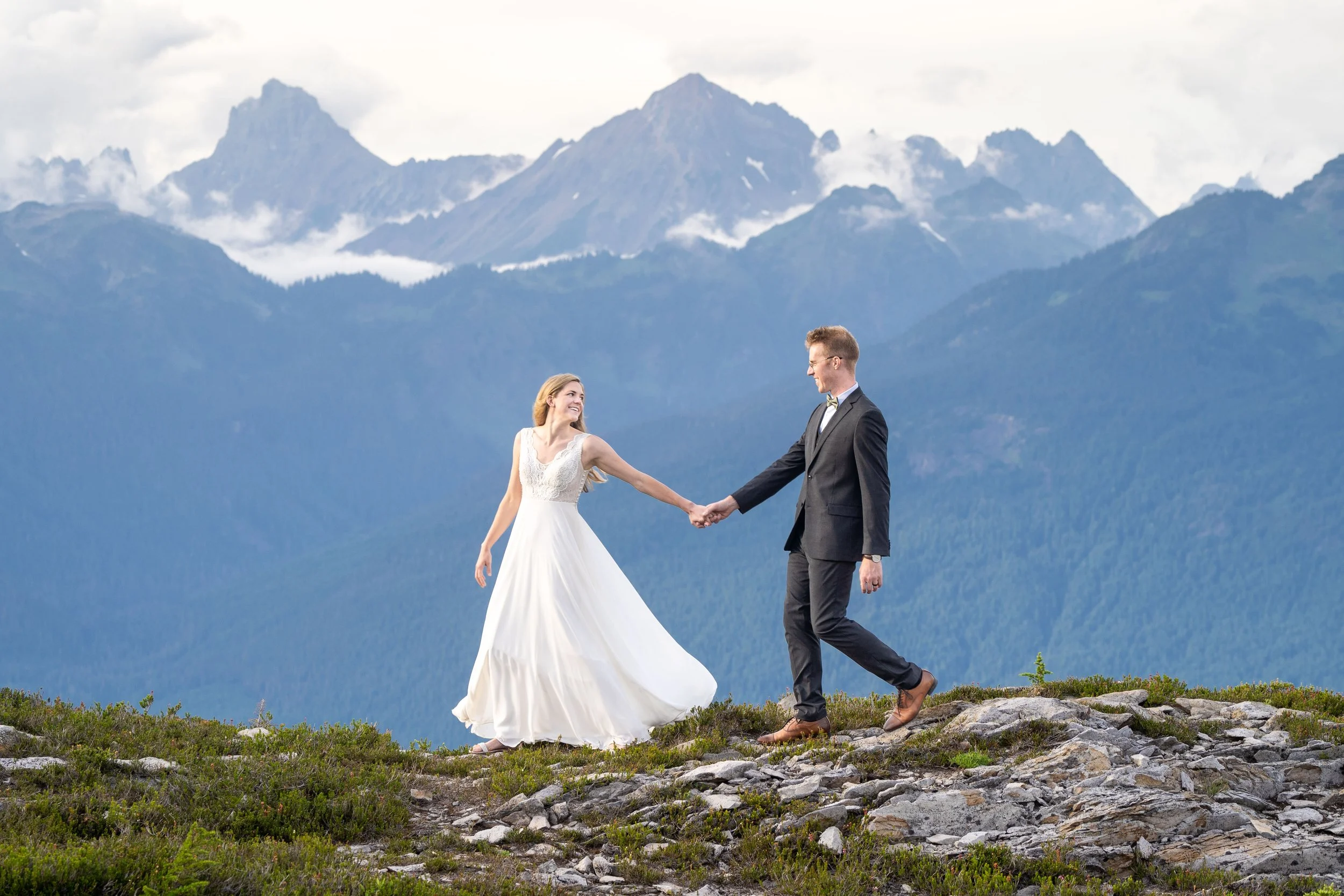 A smiling bride and groom holding hands and walking outdoors on rocky terrain with mountains and a cloudy sky in the background. The bride is wearing a wedding dress and the groom is wearing a suit.