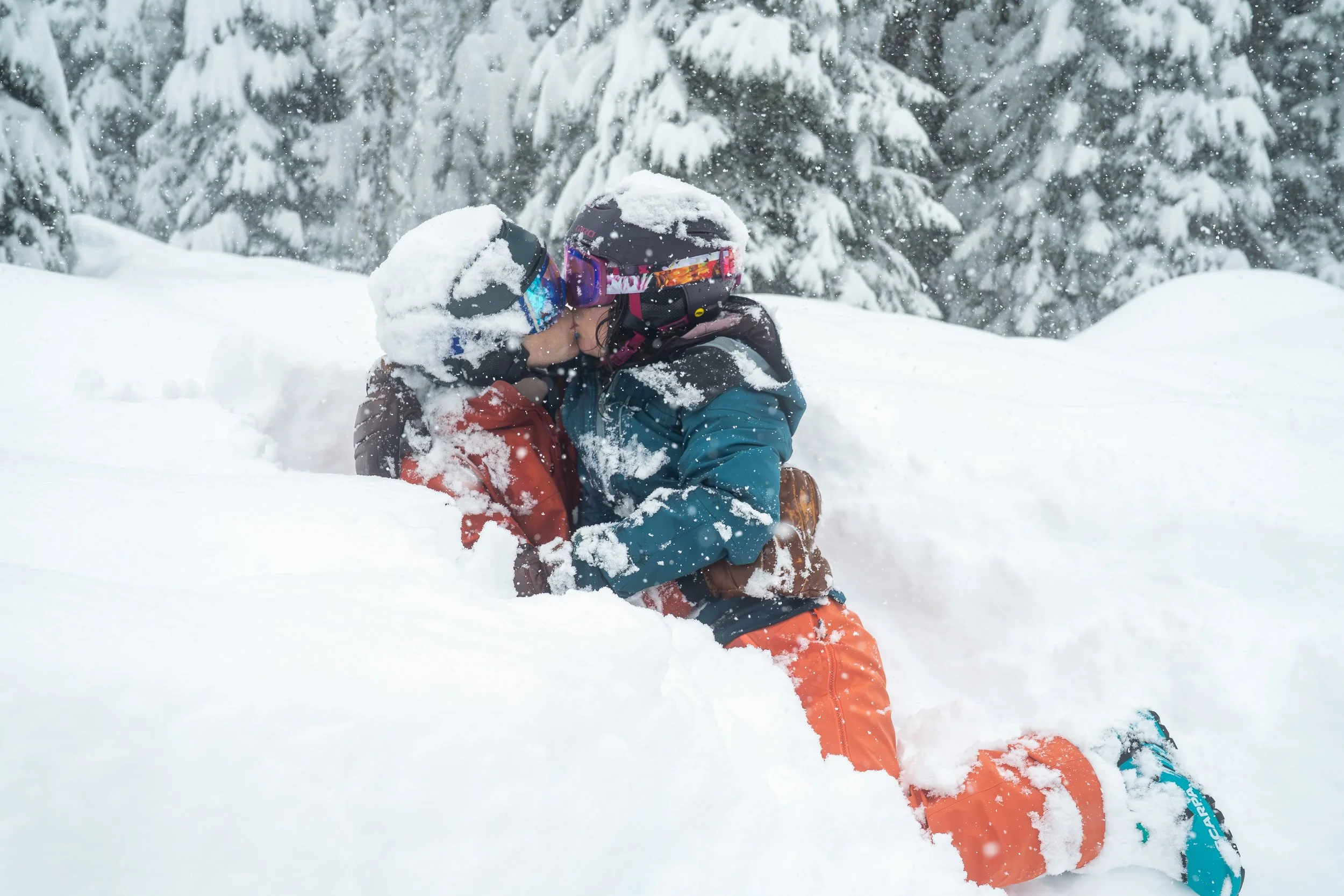 Photo of a man and woman in ski clothes, including ski goggles and helmets, kneeling in deep snow and kissing one another. It's snowing and the trees behind them are covered in snow.