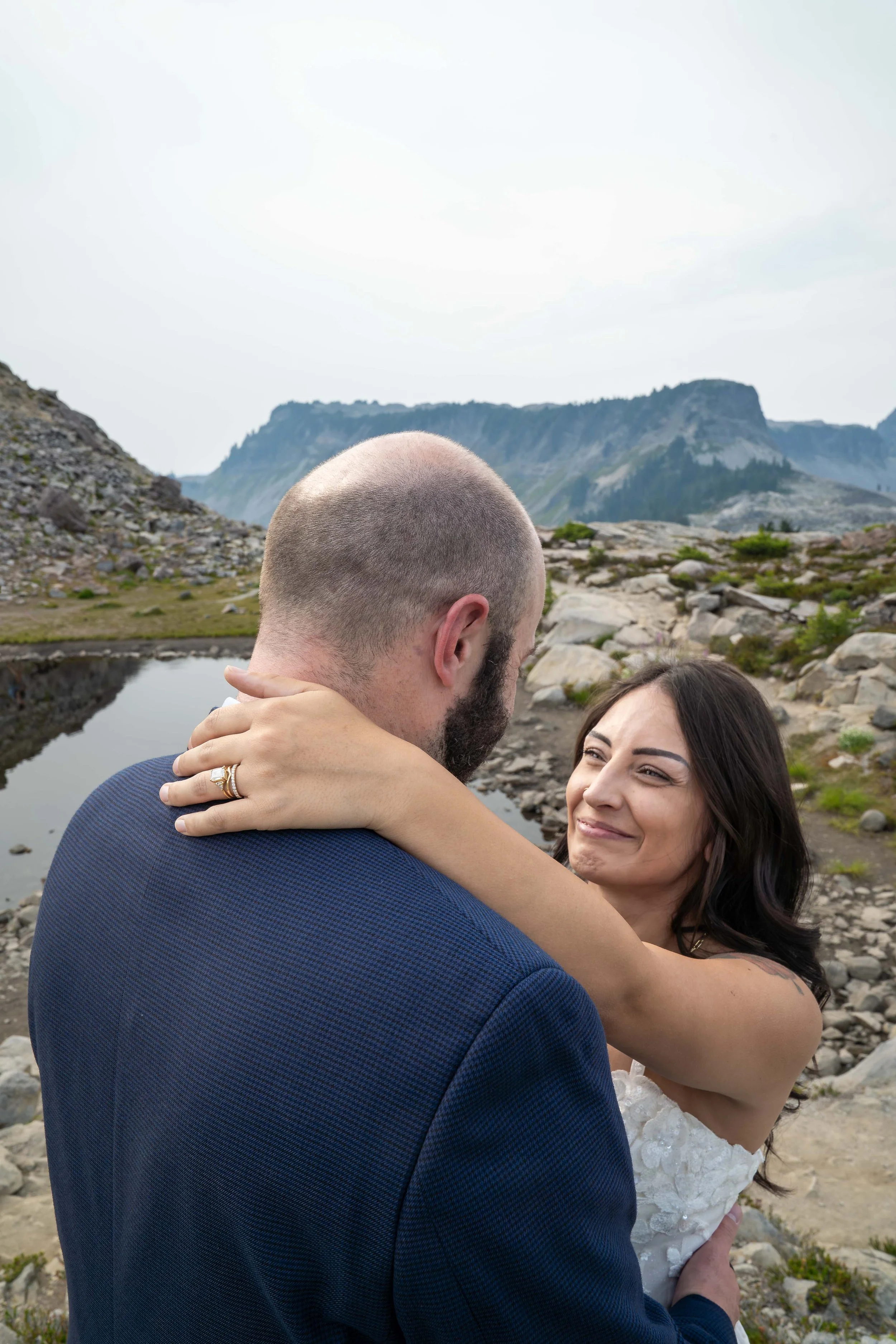 A bride and groom standing together by an alpine lake during their Artist Point elopement. The bride has her hand around her groom's neck and her wedding ring is clearly visible. Table Mountain is visible in the background.