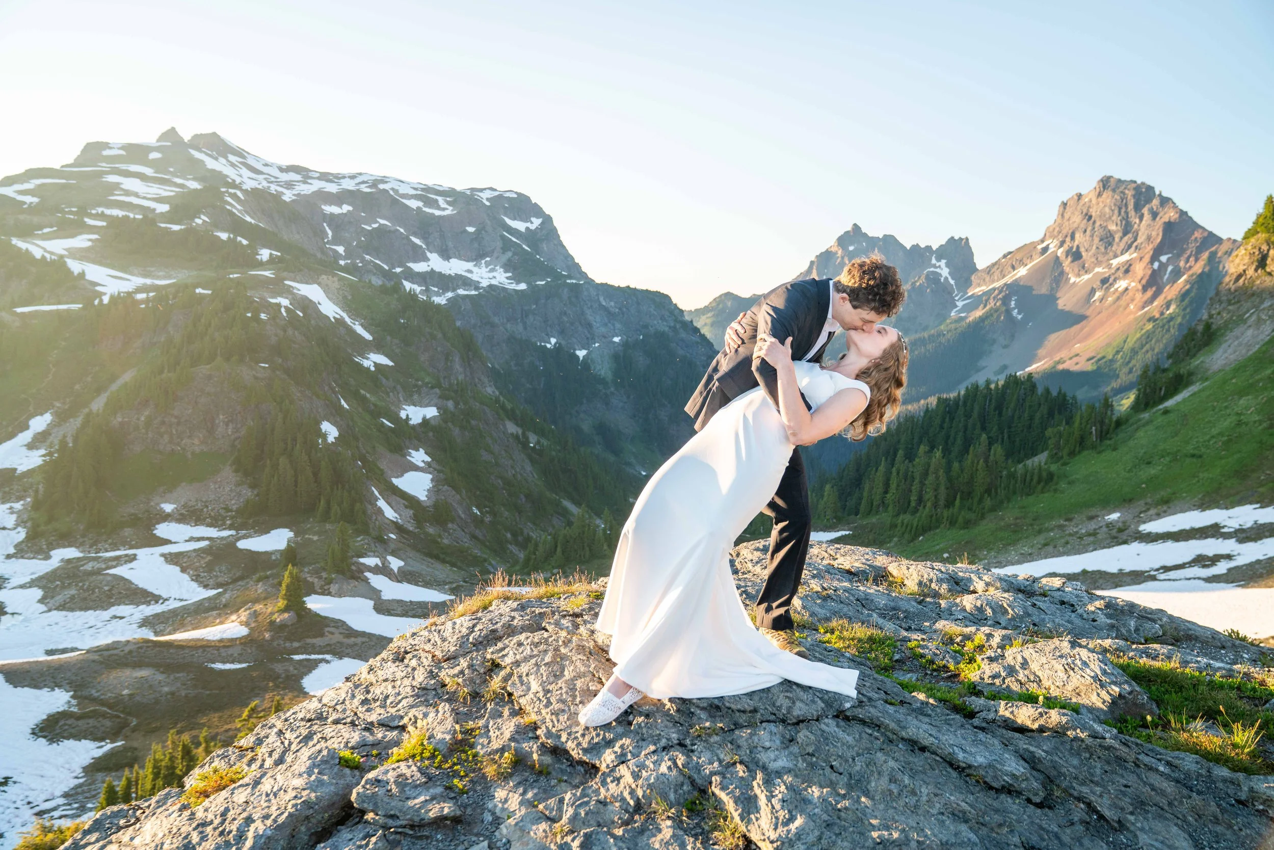 A photo of a bride and groom in wedding clothes kissing. The bride is bent over backwards and the groom is leaning down to kiss her. They're standing on a rock in the middle of an alpine meadow and there are snowy mountains in the background.