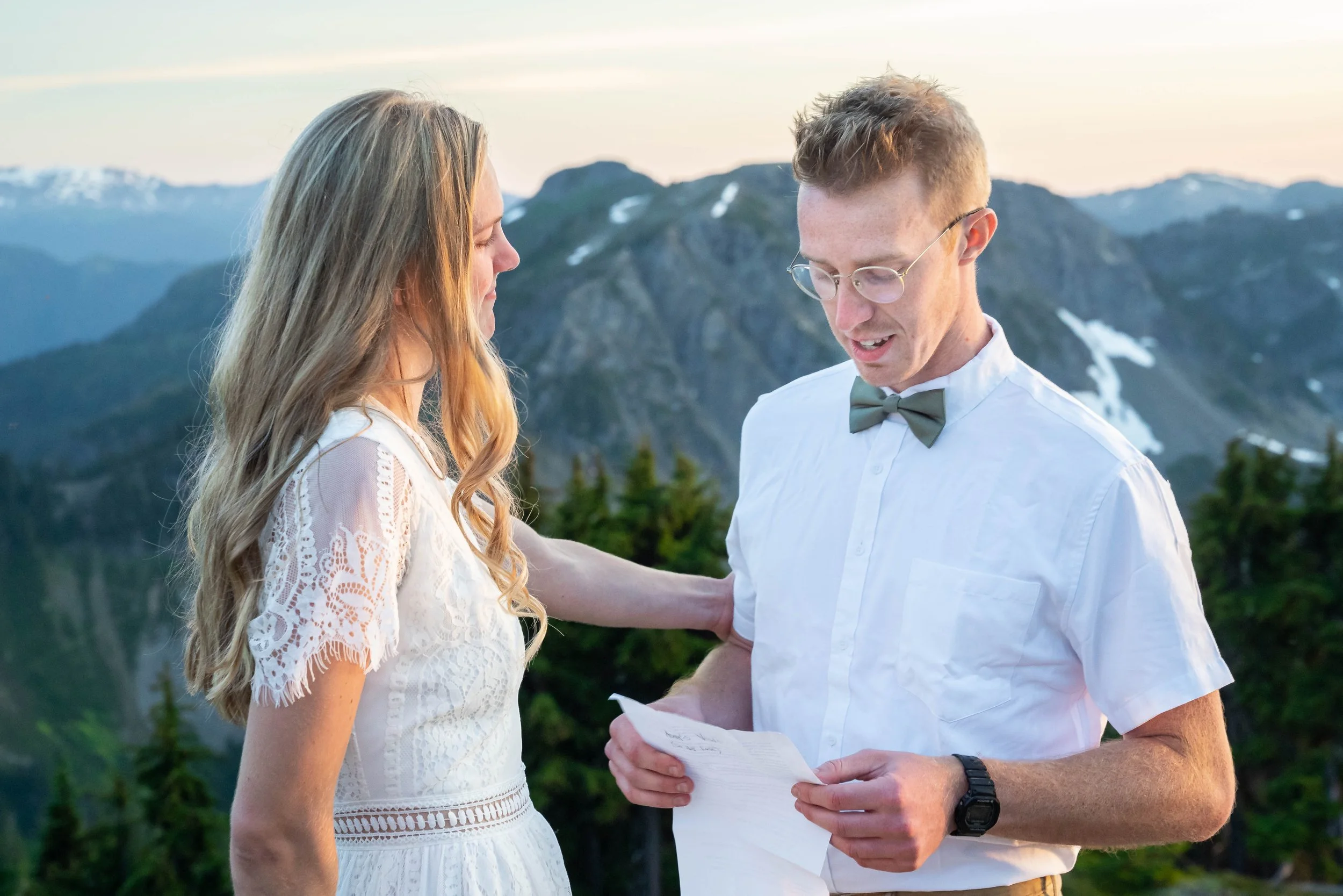 A photo of a bride and groom exchanging vows during a North Cascades elopement at sunset. The groom is reading vows from a piece of paper while his bride holds his arm, and the peaks of the North Cascades are visible behind them.