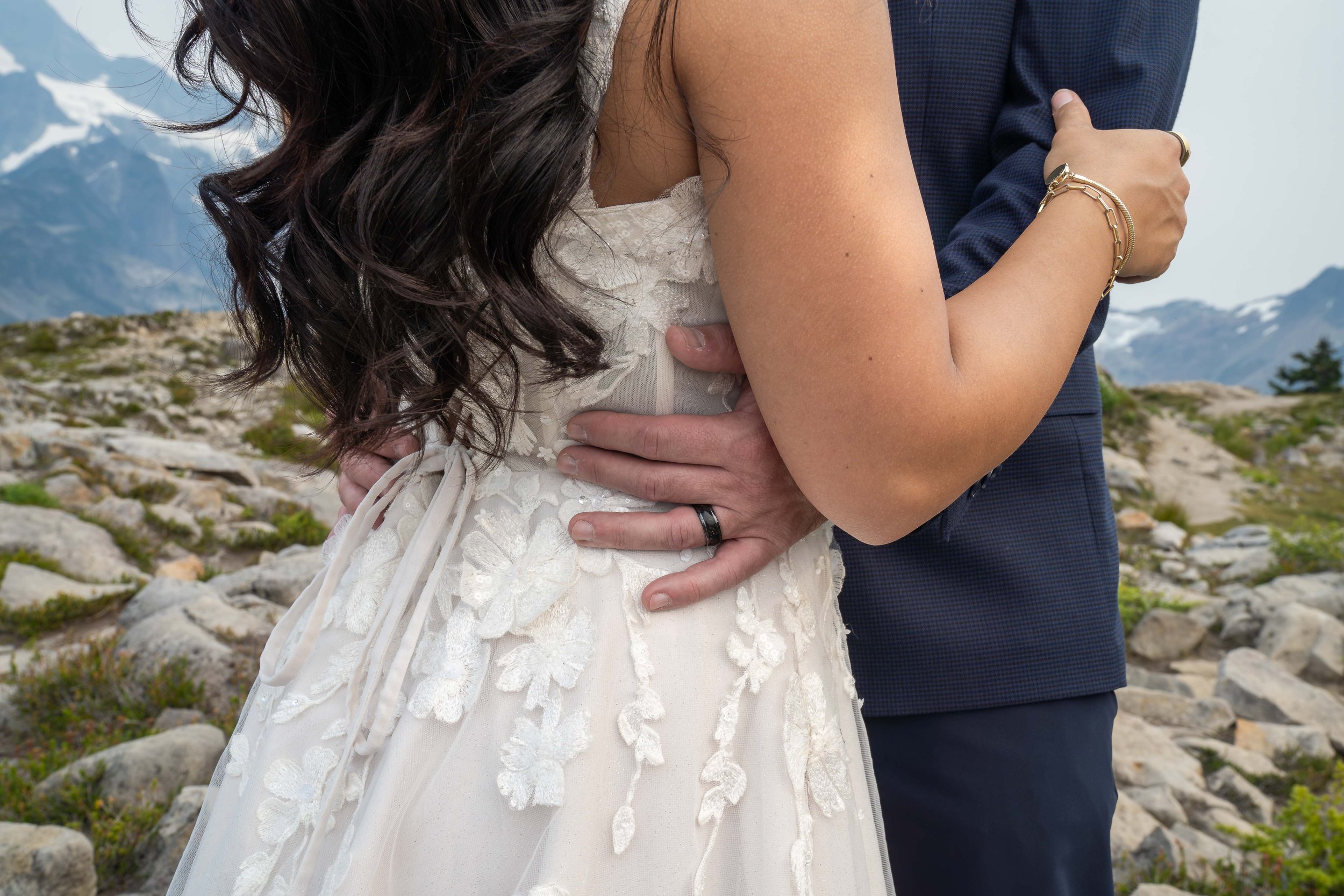 A close-up photo of a bride and groom standing together during their Artist Point adventure wedding. The groom's hands are around the bride's waist so his wedding ring is clearly visible.