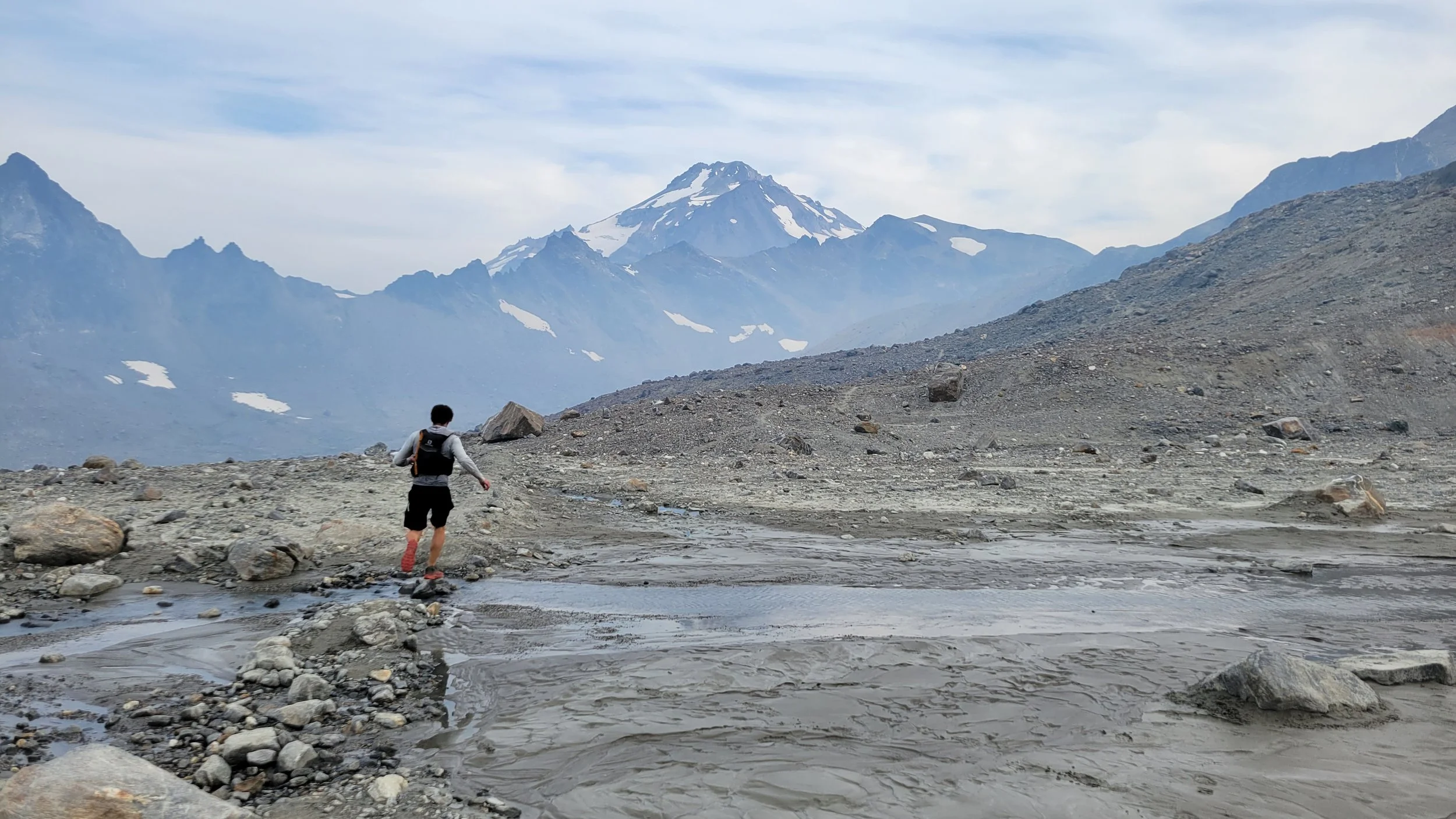 Michael, the photographer behind Wandering Sole Studios, running through a barren post-glacial landscape around Glacier Peak, in Washington. He is running away from the camera and Glacier Peak is visible in front of him.