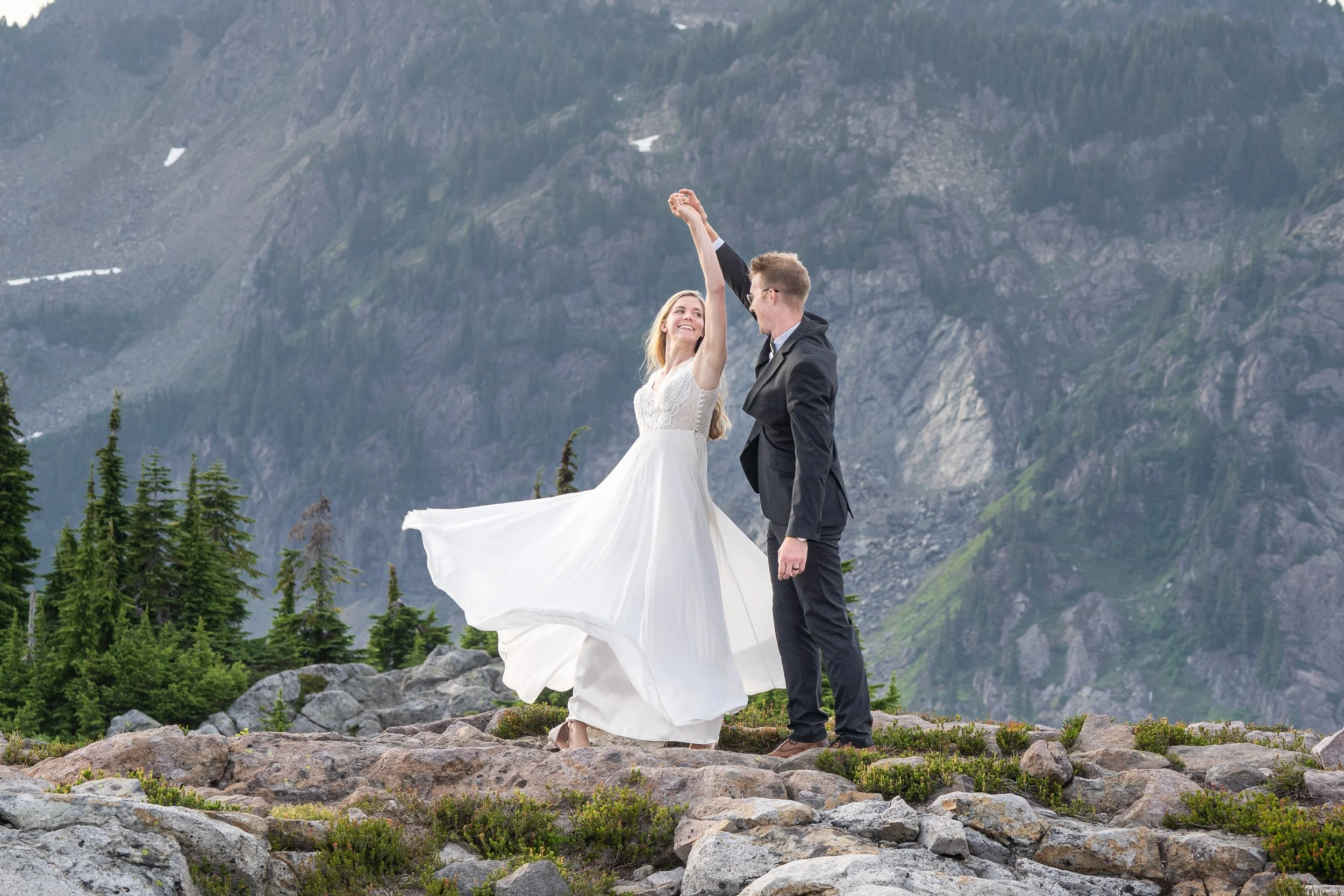 Photo of a bride and groom in wedding clothes dancing together on rocky alpine terrain during an Artist Point elopement. The bride is spinning and her wedding dress is swirling in the air.