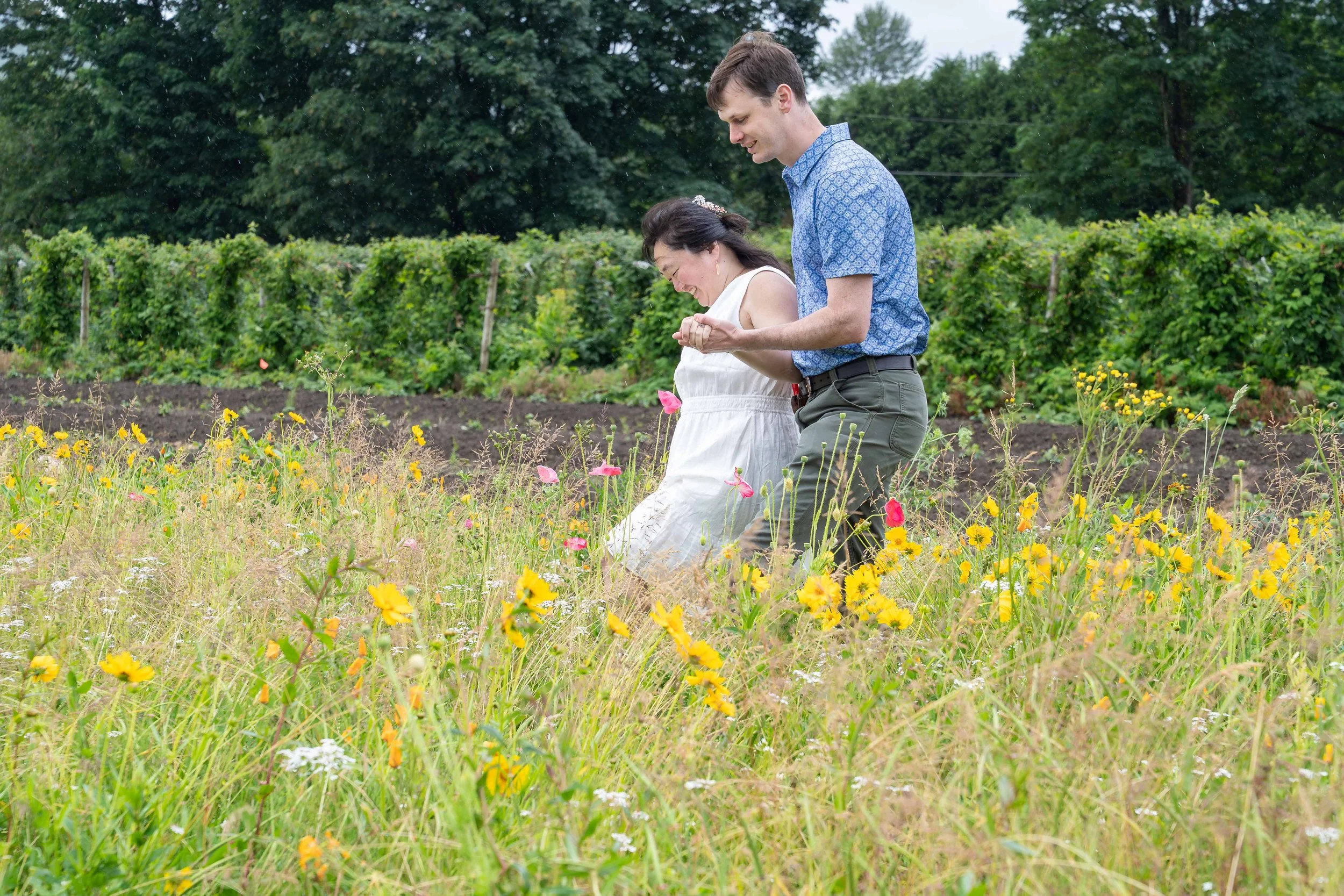 A couple holding hands and walking through a field of yellow flowers during a Bellingham elopement photography session. There are raspberry bushes and trees in the background.