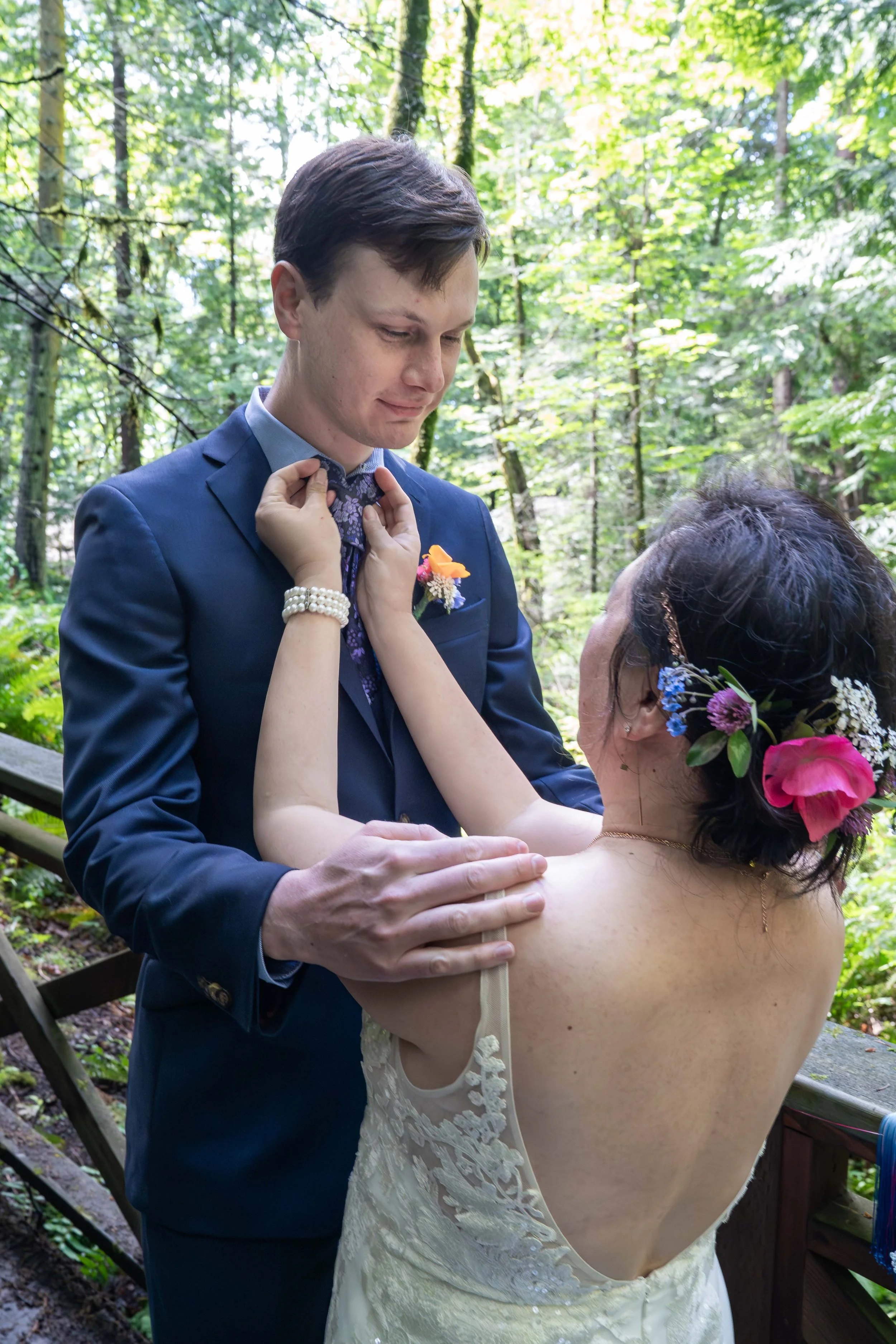 A bride reaches up to adjust her groom's tie after a first look in Larrabee State Park. The groom is looking down at the bride and holding her shoulders, and they're surrounded by lush forest.