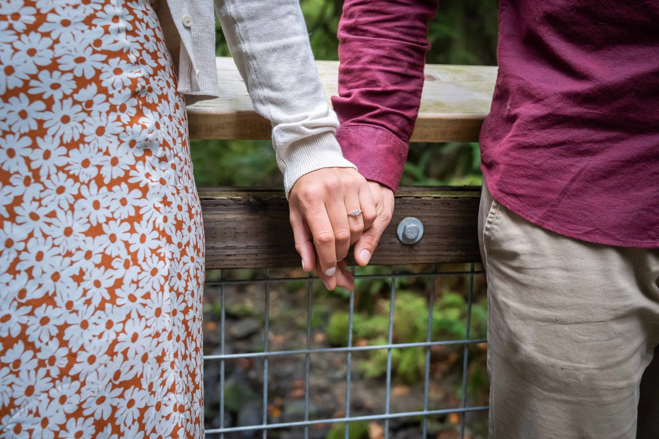 A close-up photo of a couple from their knees to their chests. They're holding hands and the woman's engagement ring is in the center of the photo. They're on a wooden bridge in the middle of a green forest.