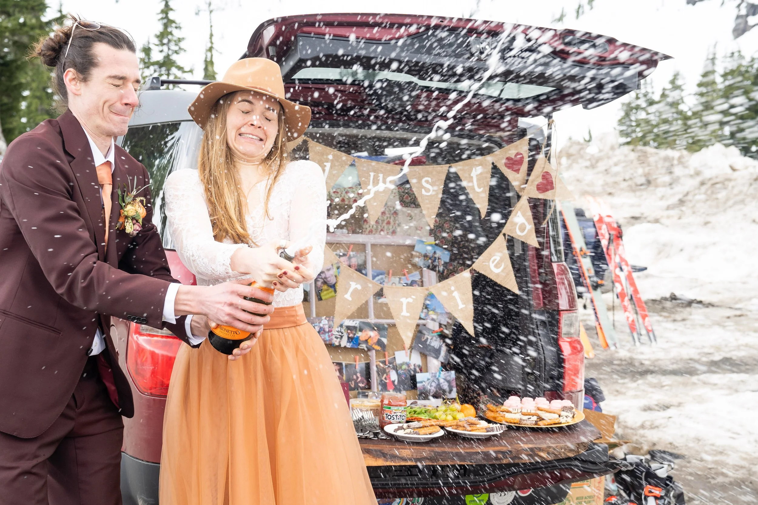 A photo of a bride and groom popping a bottle of champagne. Champagne is exploding out of the bottle as they both have their eyes closed. Behind them is a sign that says Just Married with a platter of desserts.