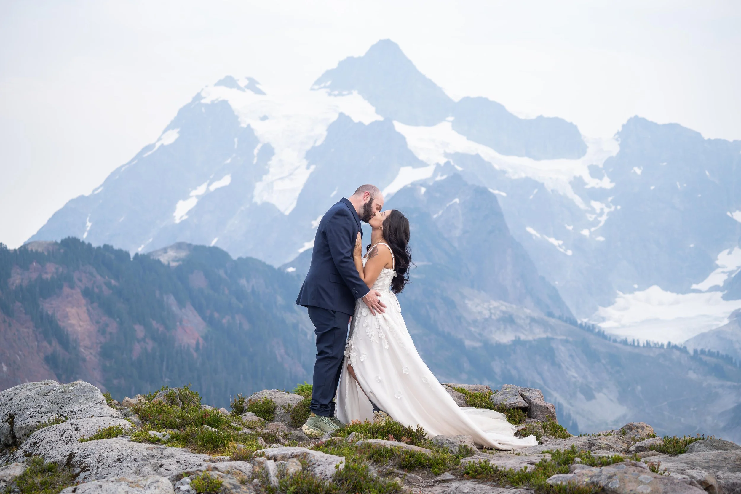 A bride and groom in wedding clothes kissing in front of Mt. Shuksan during their Artist Point elopement. They are standing in a rocky alpine meadow and the mountain is hazy from smoke, but still clearly visible.