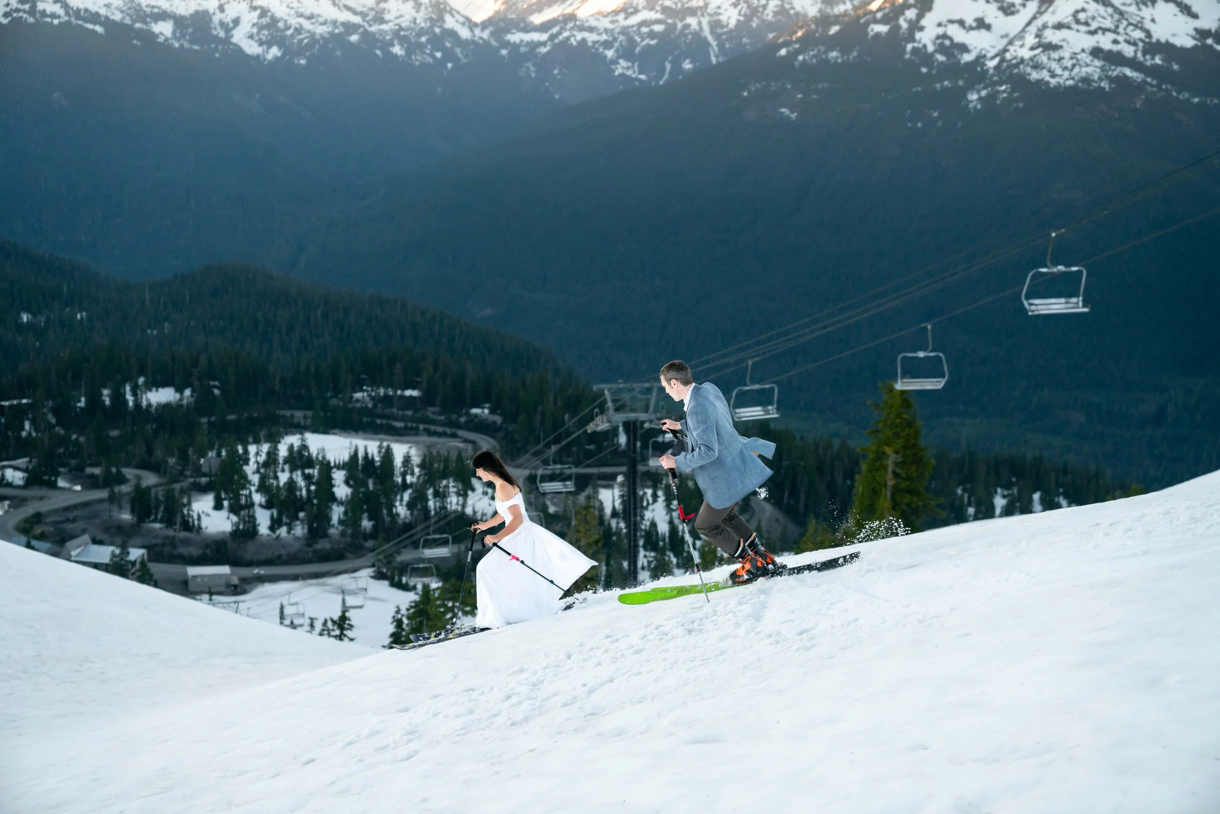 A photo of a man and woman in a suit and wedding dress skiing down a snowy slope with mountains in the background.