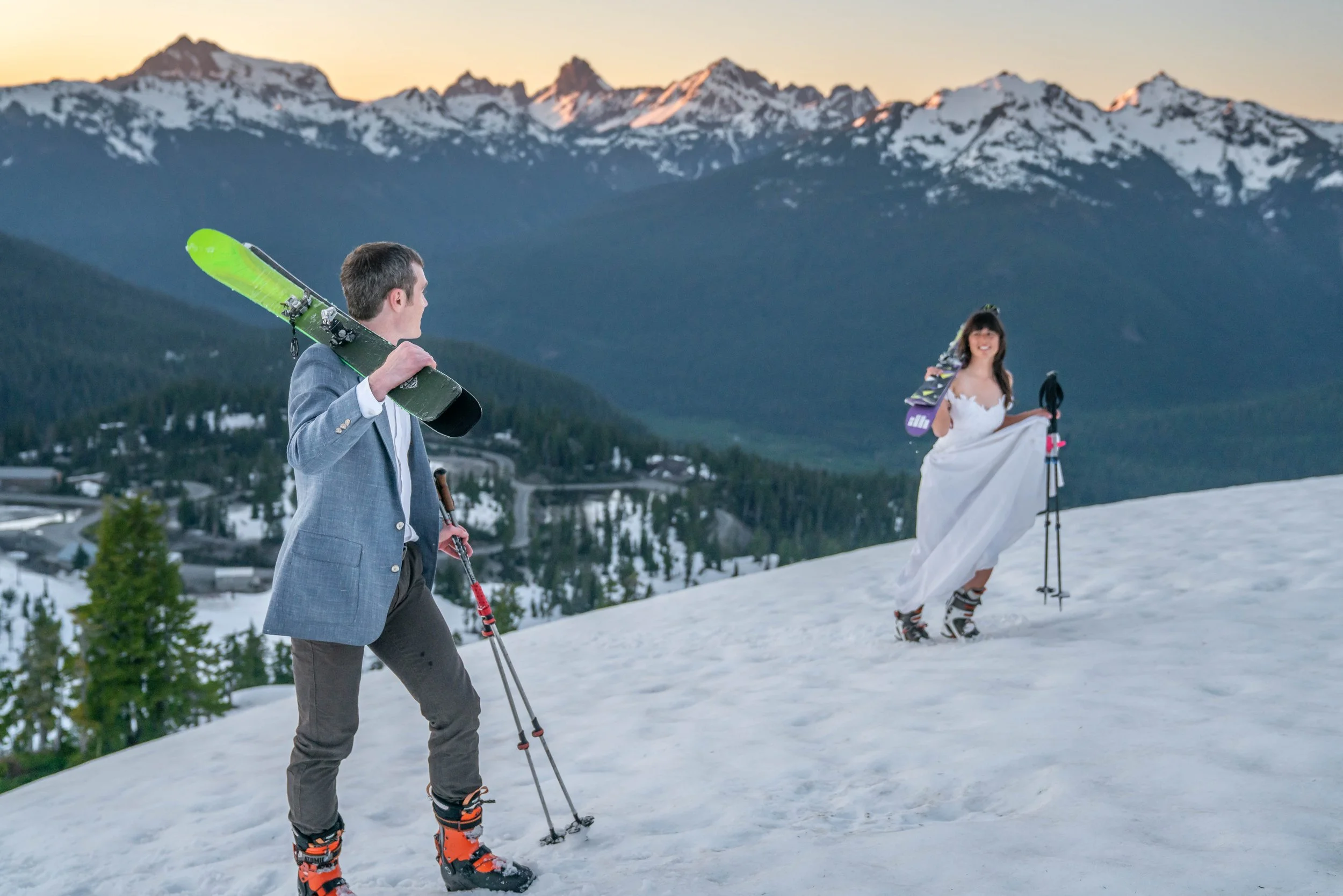 A photo of a couple in wedding clothes and ski boots walking uphill while holding skis over their shoulders. They're on snow and snowy mountains at sunset are visible behind them.