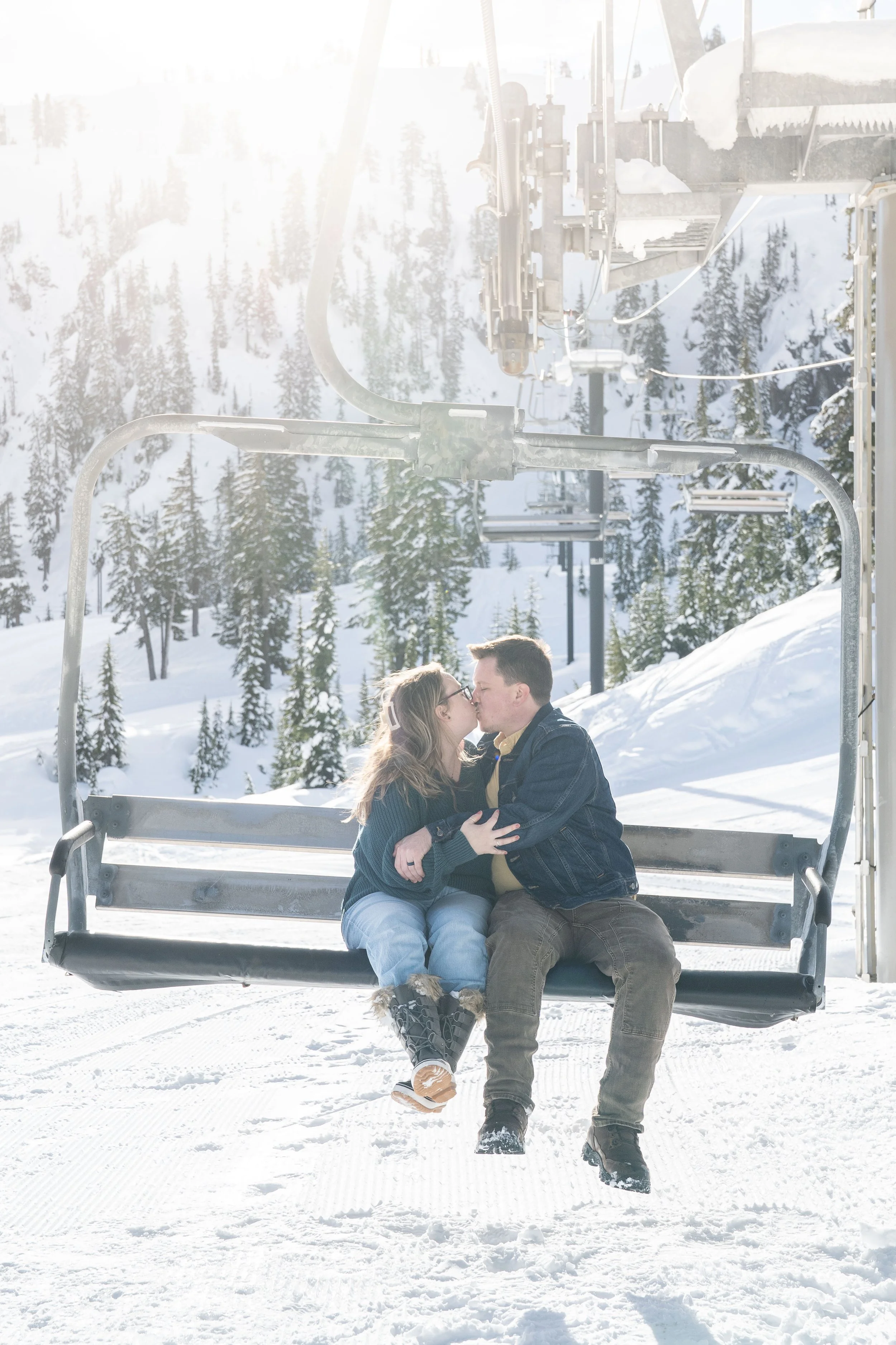 A photo of a couple holding each other and kissing on a ski lift chair at the Mt Baker Ski Area. It's snowy and the sun is shining.