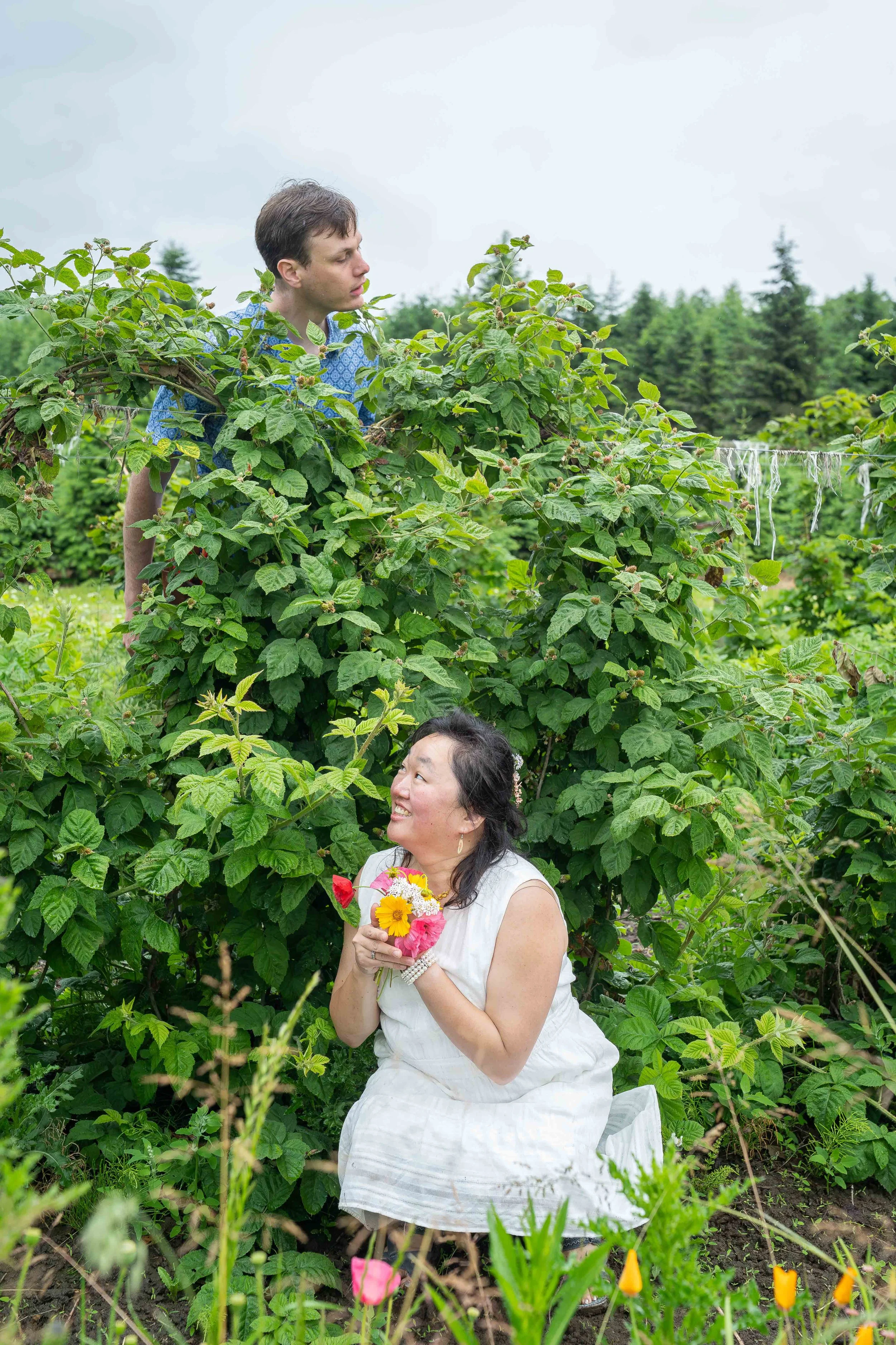 A playful engagement-style photo of a bride holding a bouquet and hiding beneath a tall raspberry bush, while her groom peers over the top of the bush looking for her.