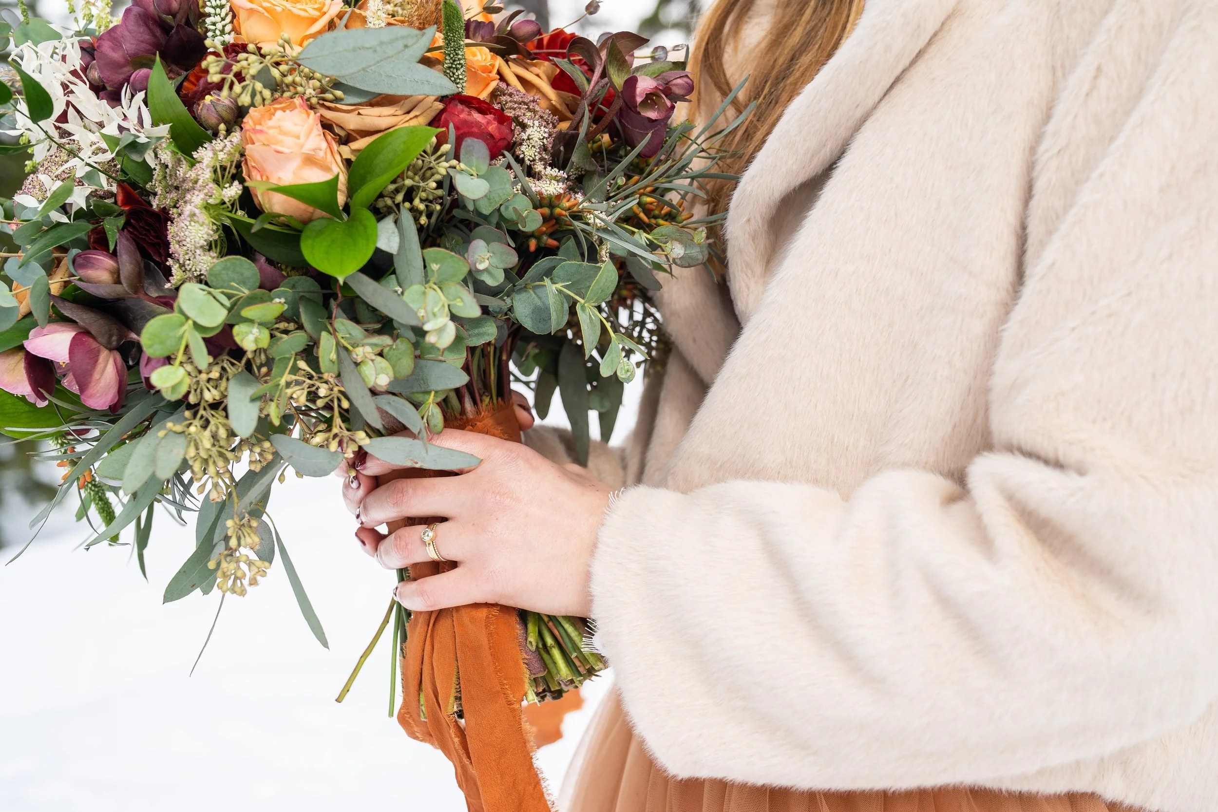 A photo of a bride holding a bouquet of flowers. The bride is wearing an orange wedding dress and white fur coat, and her wedding ring is visible on her finger.