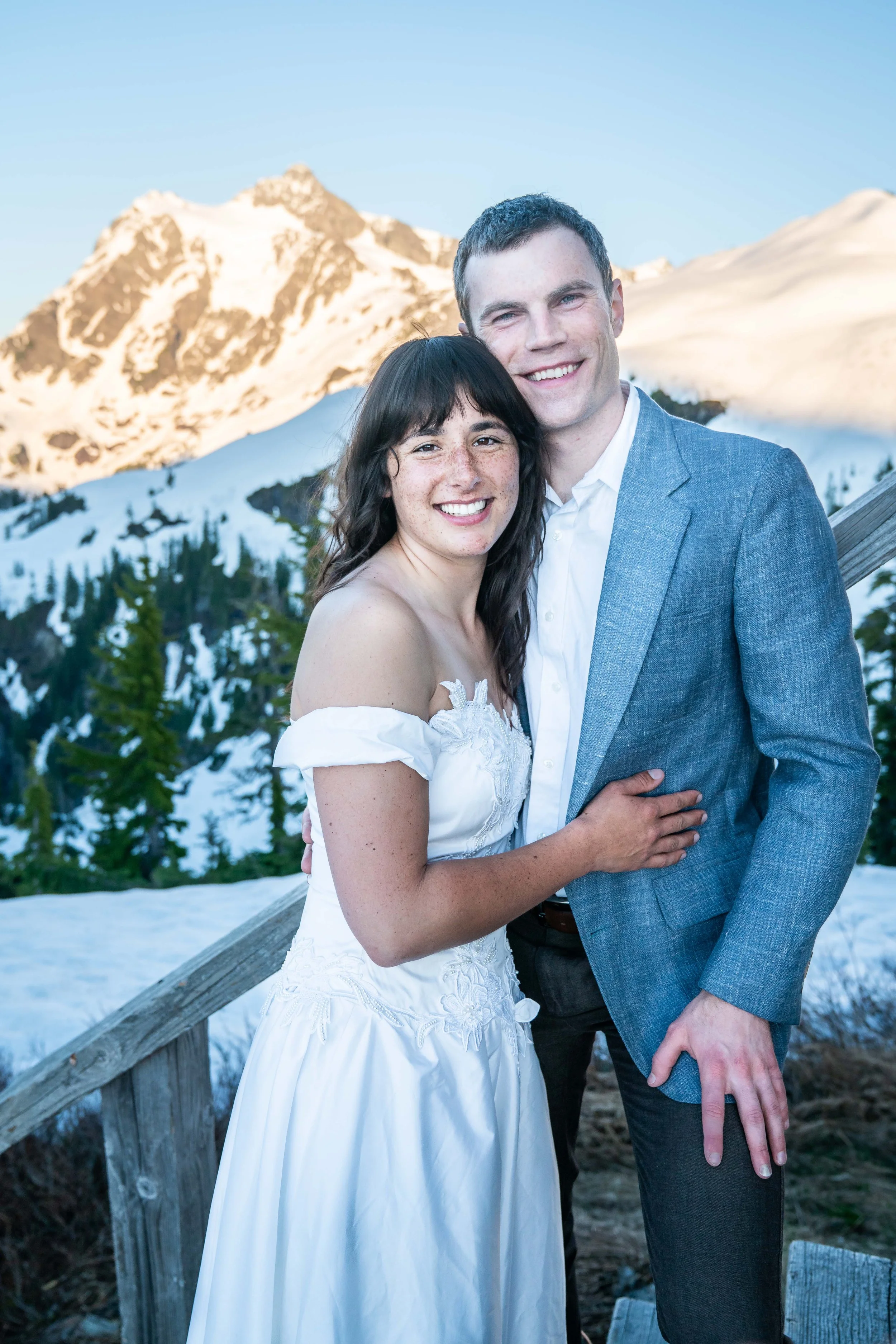 A photo of a couple in a suit and wedding dress standing together and smiling at the camera. There's snow around them and Mt. Shuksan in the background.