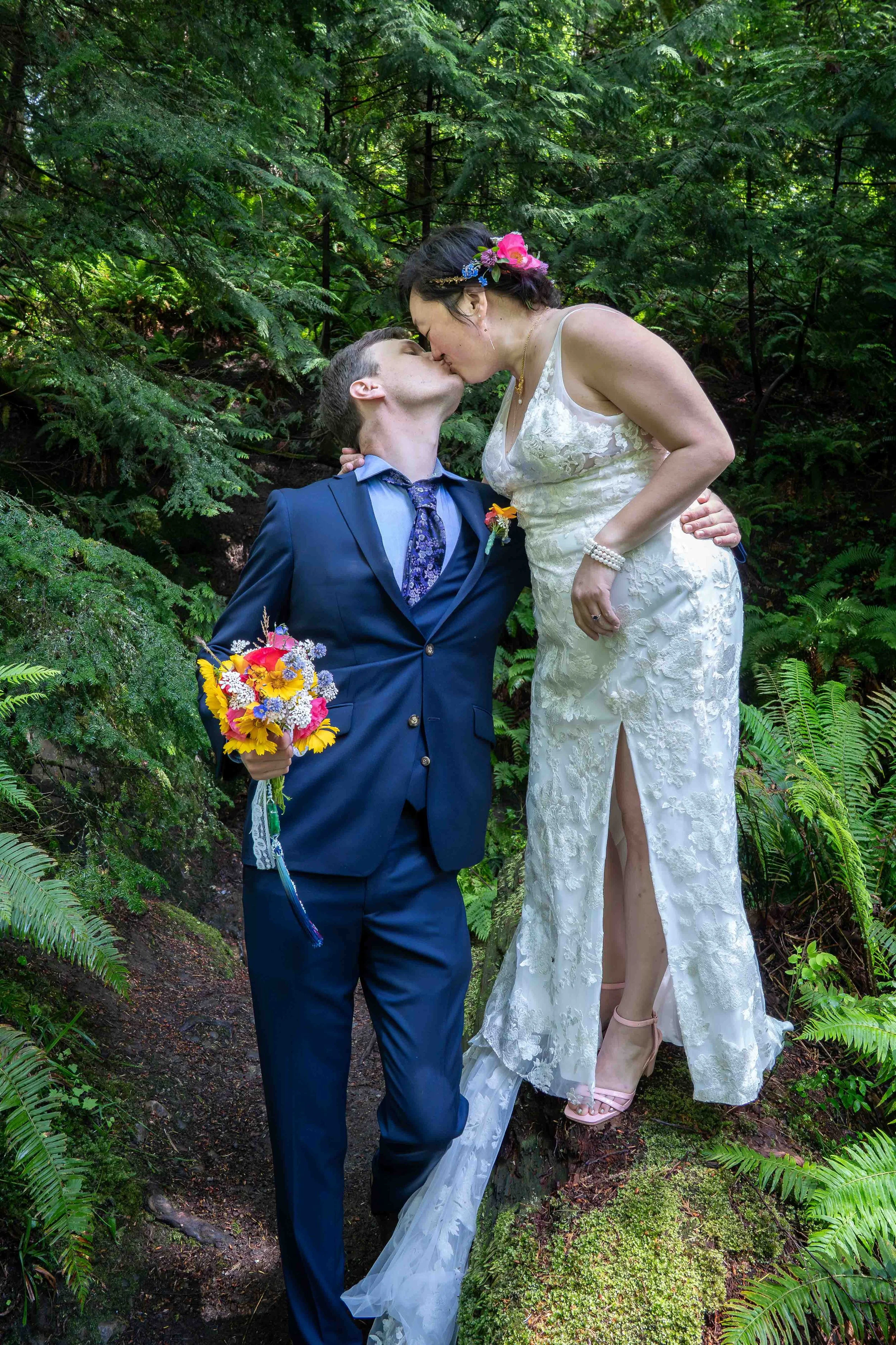 A bride and groom kiss after their first look during a Larrabee State Park elopement. The bride is standing on a log and leaning down to kiss the groom.