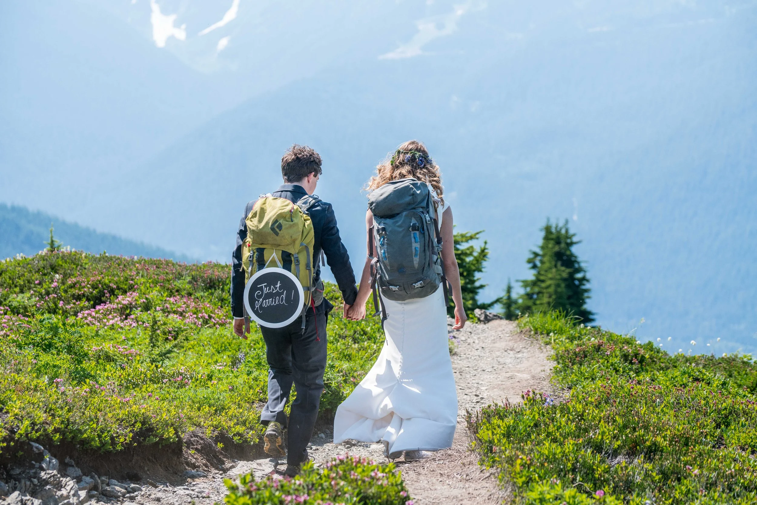 Photo of a couple in wedding clothes walking down a trail, with backpacks on and a hanging sign that says "Just Married".