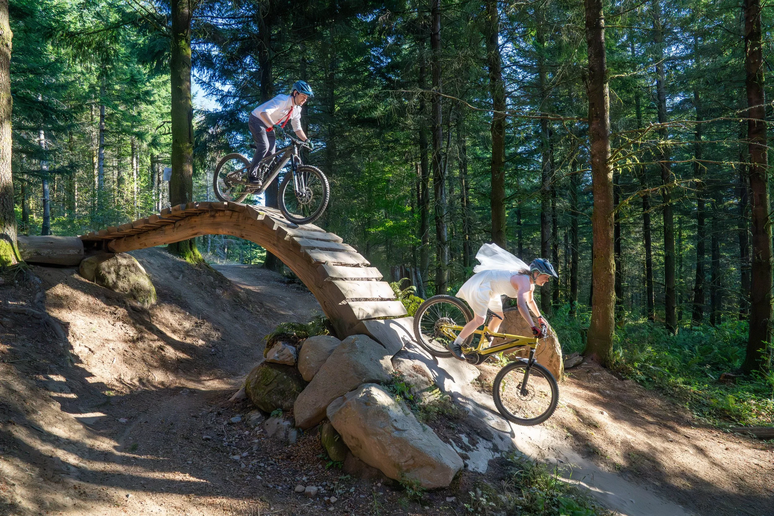 Photo of a mountain biking elopement on Unemployment Line wooden bridge roll on Galbraith Mountain in Bellingham. The bride is at the bottom of the roll wearing a wedding dress with cut-off legs and the groom is right behind her in a suit and tie.