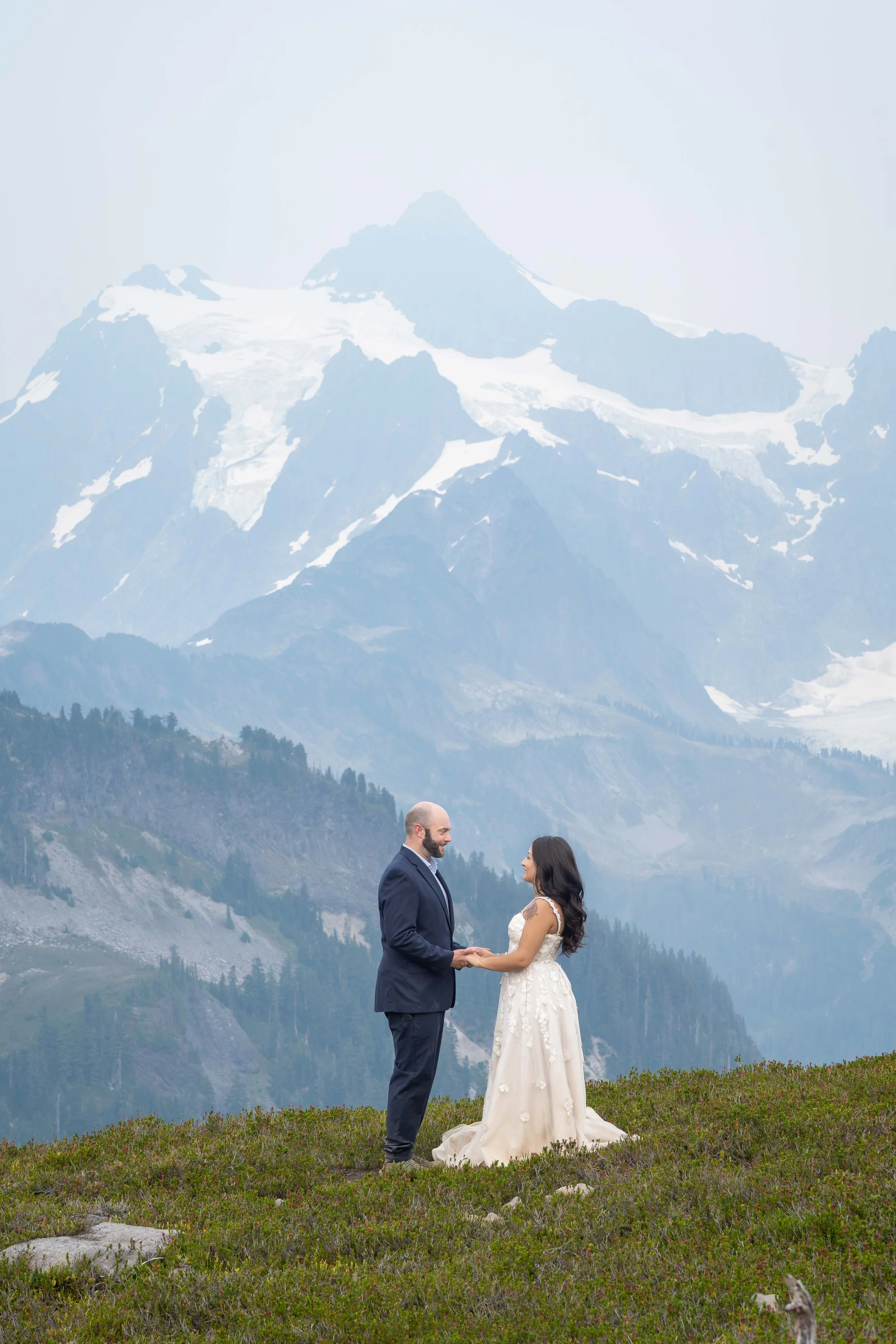 A bride and groom in wedding clothes stand together and hold hands in a heather meadow on Ptarmigan Ridge during their Mt. Baker Elopement. Mt. Shuksan is visible directly behind them, but it's hazy due to wildfire smoke.