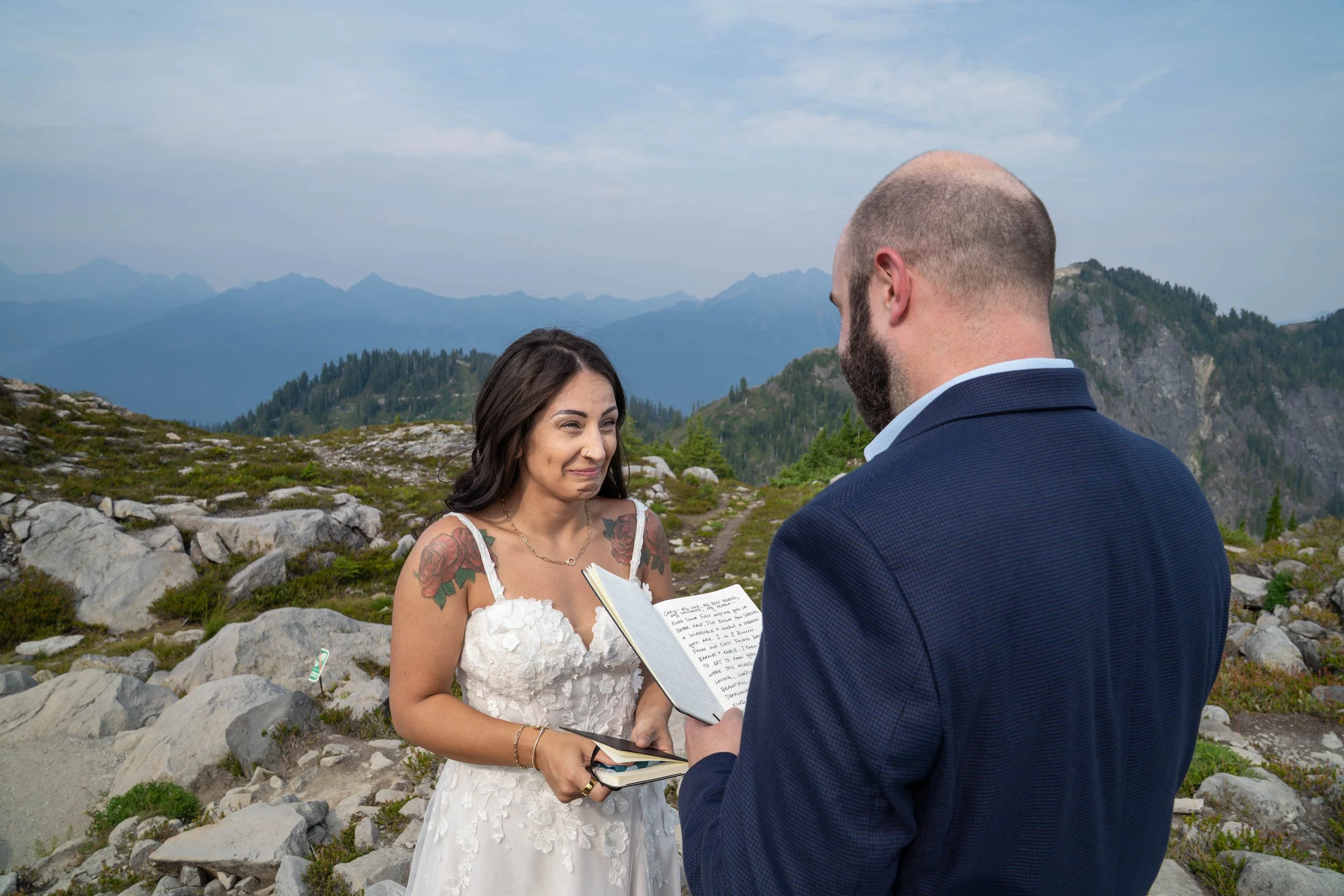 A groom reading his handwritten vows from a vow book during an elopement ceremony at Artist Point, near Mt Baker while his bride holds back tears. They are standing in an alpine meadow with the North Cascades visible behind them.
