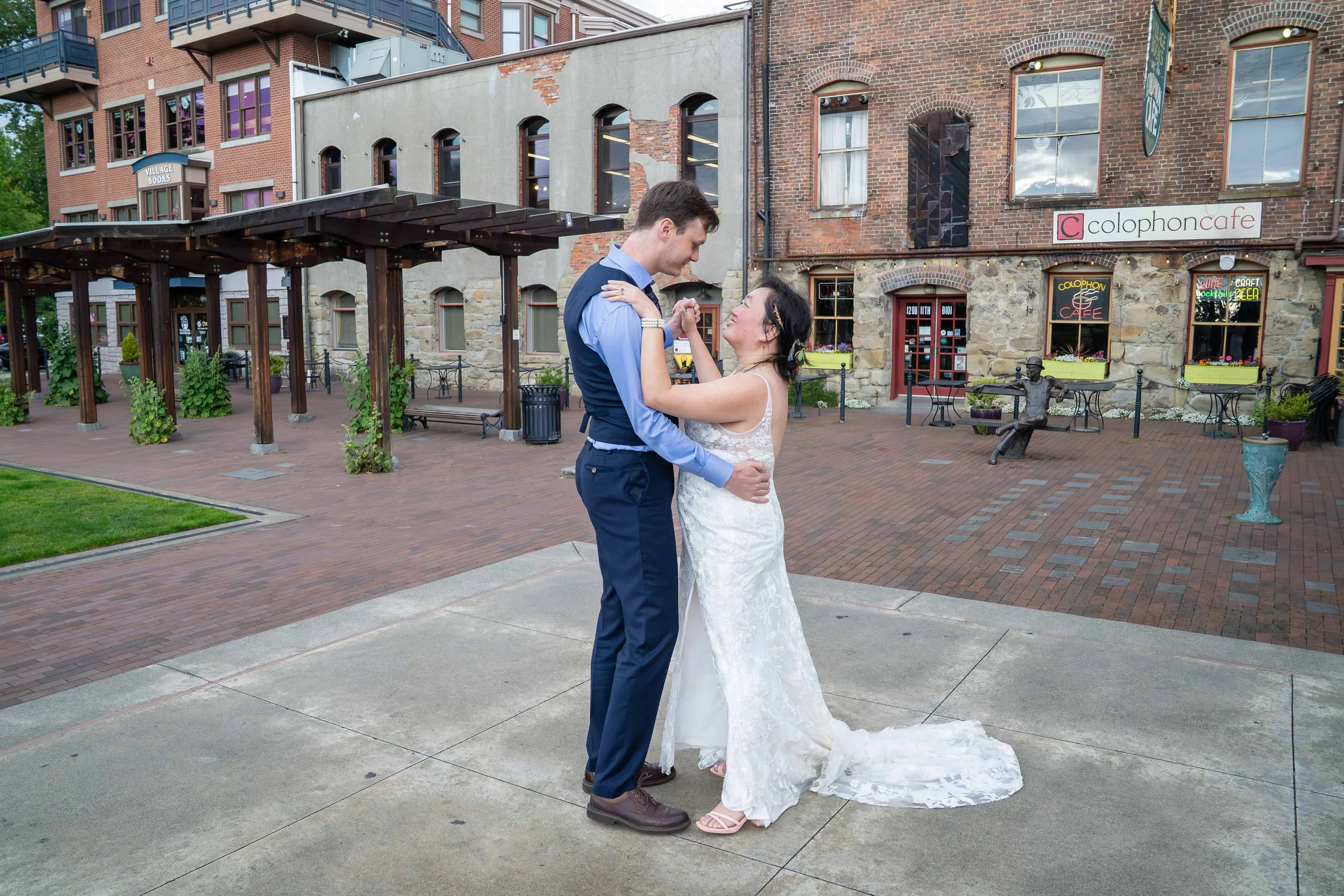 A bride and groom hold their first dance on the Village Green in historic downtown Fairhaven to celebrate their Bellingham elopement. They are slow dancing together and smiling at one another, with a brick-lined plaza and old stone buildings behind.