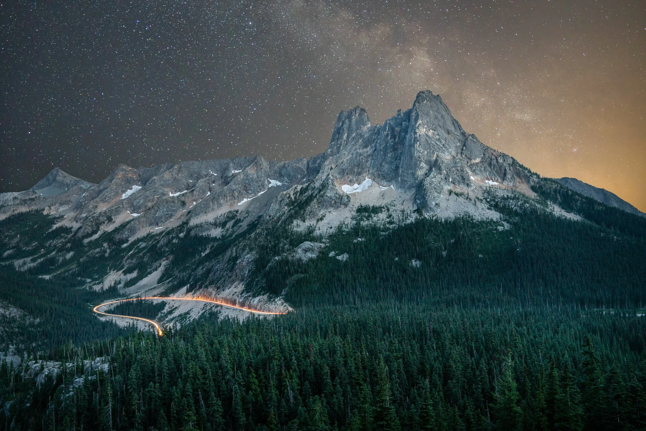 A landscape astrophotography image of Washington Pass with the Liberty Bell group and the Milky Way in the sky behind it. A car is going around the hairpin turn on Highway 20, lighting up the road.