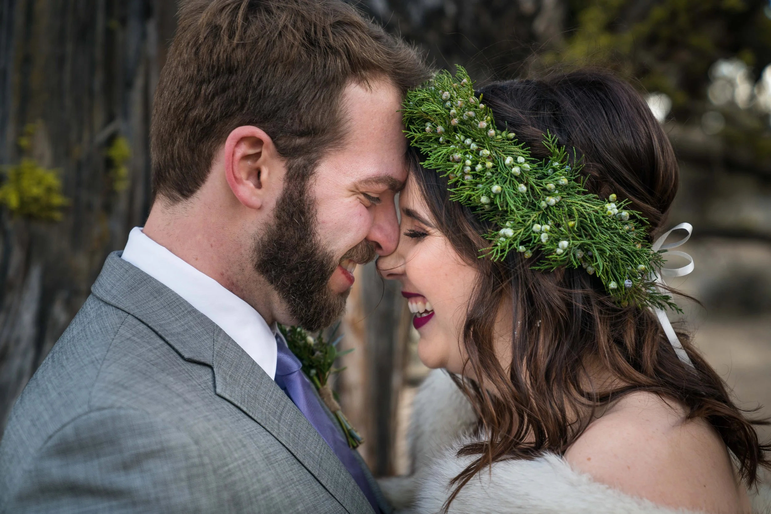 A couple in wedding clothes smiling and pressing their foreheads together. The bride has a headband made of juniper and they are standing in front of a mossy tree.