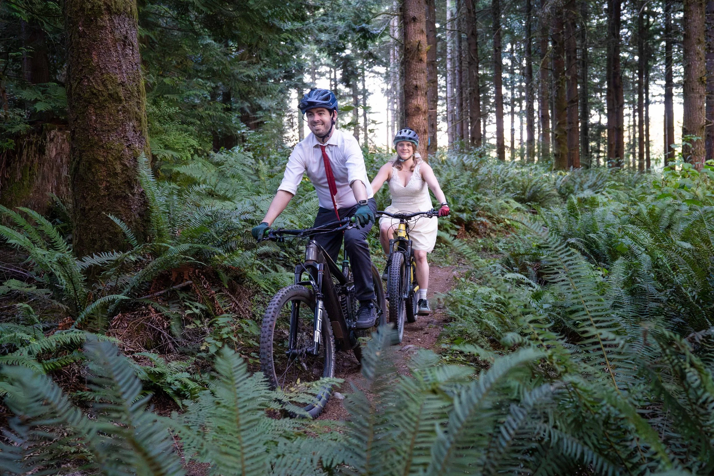 A couple in wedding clothes riding mountain bikes on singletrack through lush ferns and mossy trees during their mountain bike elopement on the 187 trail on Galbraith Mountain, in Bellingham, Washington.