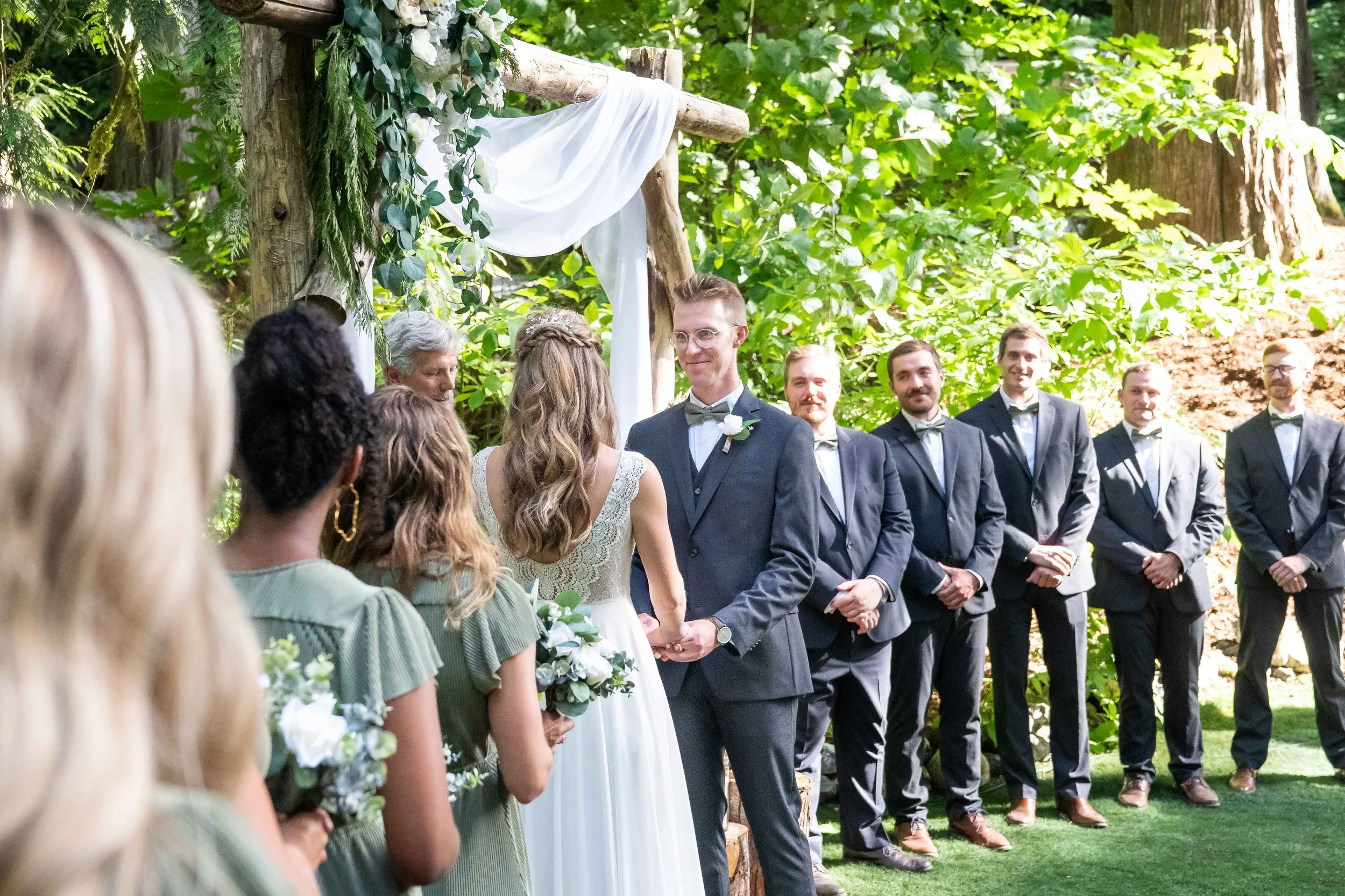 Photo of a wedding ceremony in the woods at Creekside Celebrations in Skagit County, Washington. The bride is reading her vows while the groom smiles at her and the groomsmen look on.